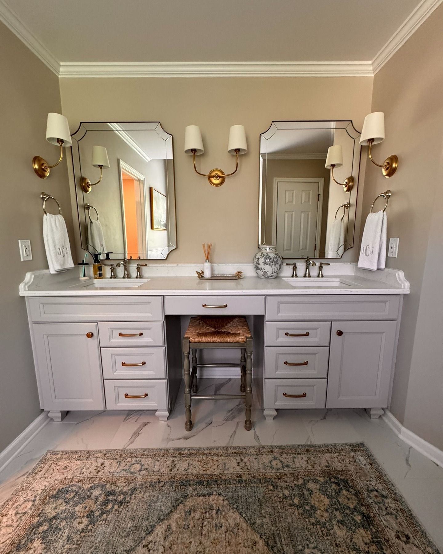 Bathroom with light gray vanity, marble countertop, two mirrors, gold sconces, and a patterned rug.