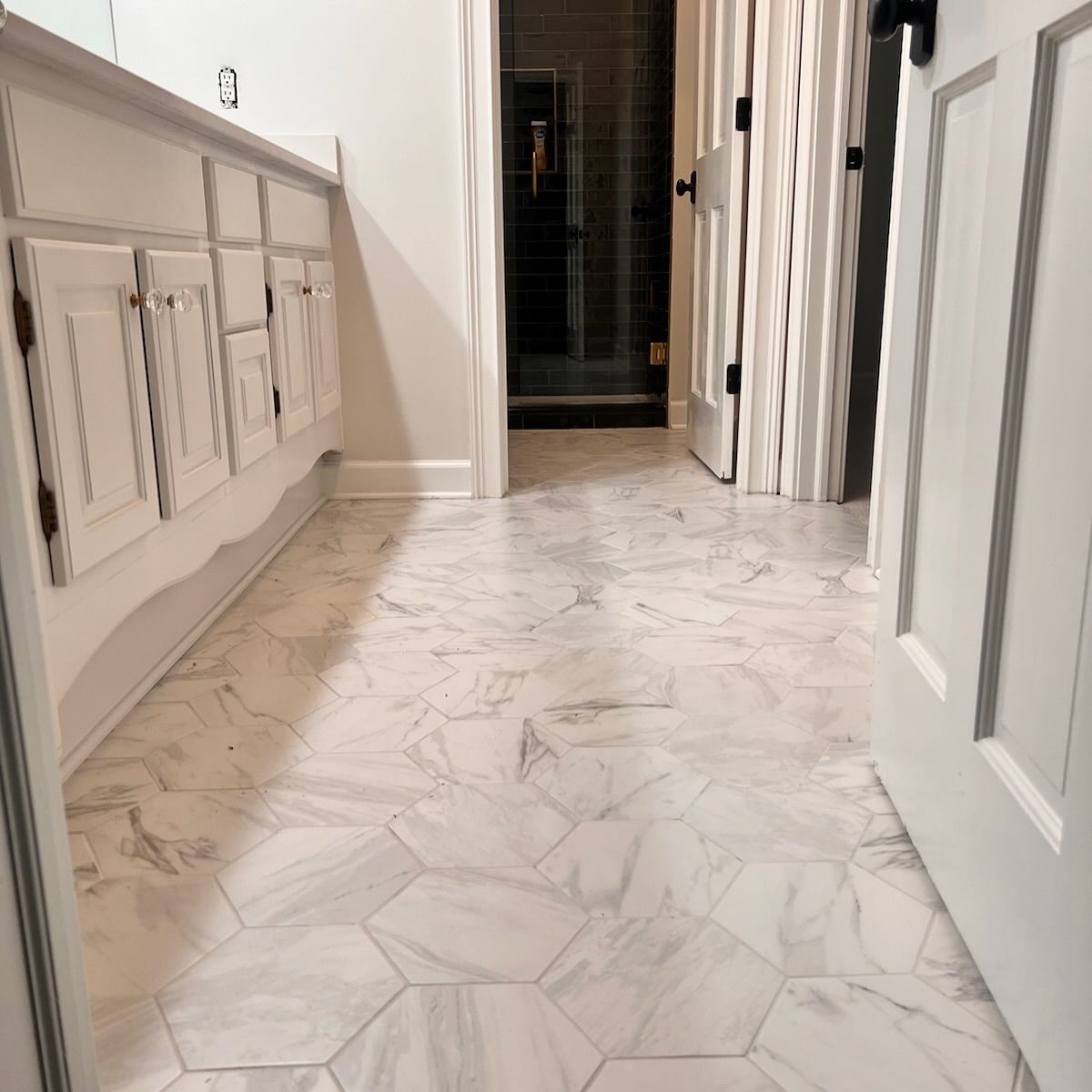 Bathroom with white hexagon-patterned floor tiles and white cabinets. Doorways lead to a shower and another room.