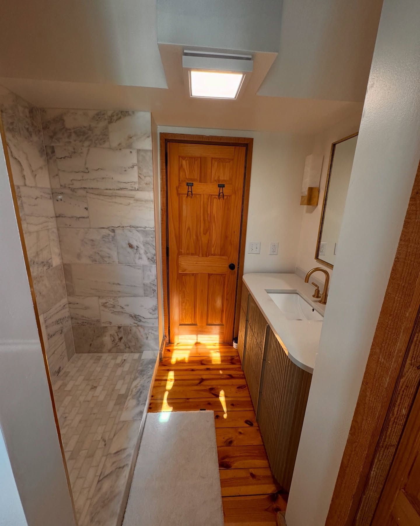Narrow bathroom with marble shower, wooden door, long vanity, and natural light.