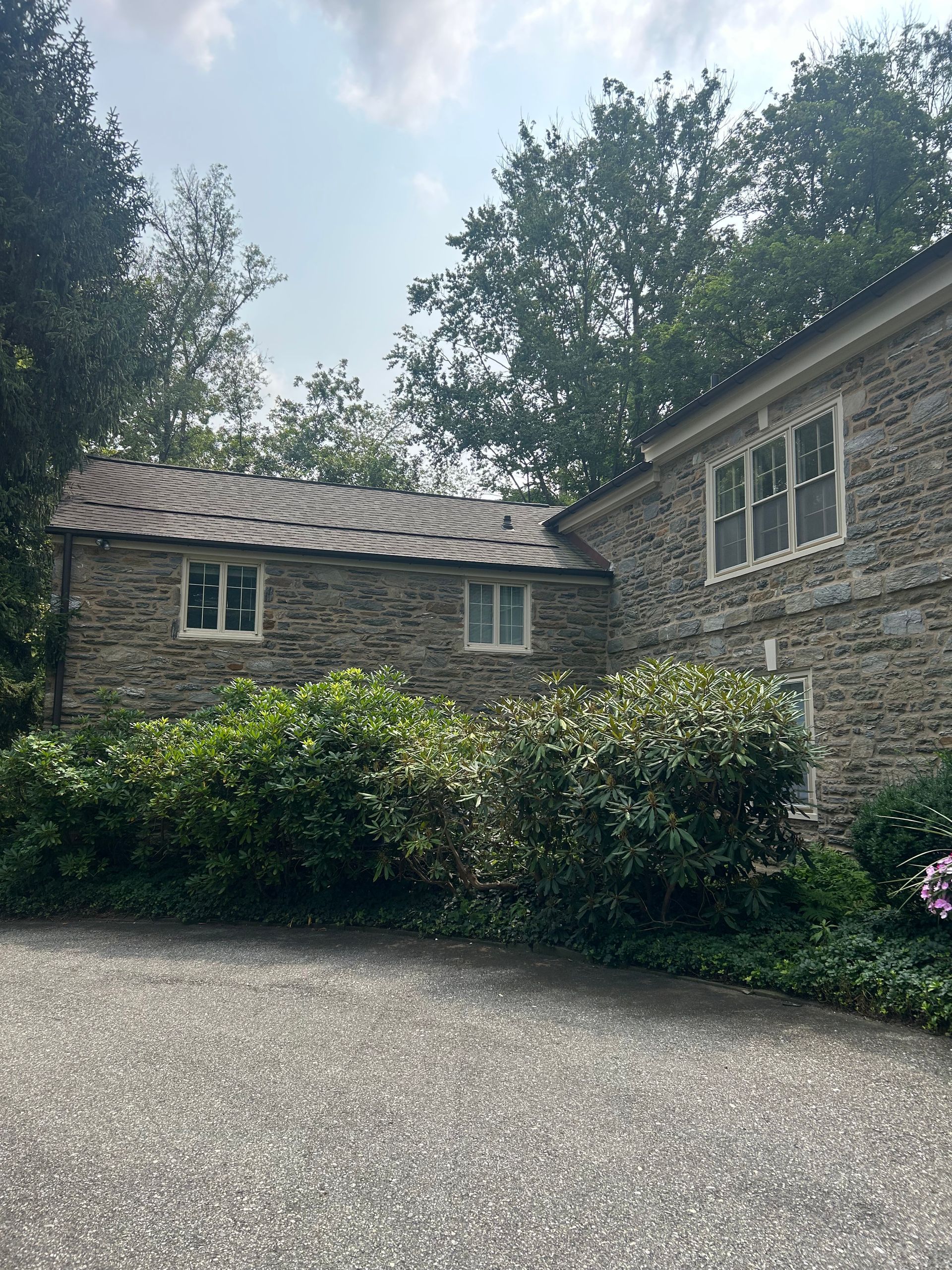 Stone building with two sections, windows, and a dark shingled roof, surrounded by green bushes and trees.