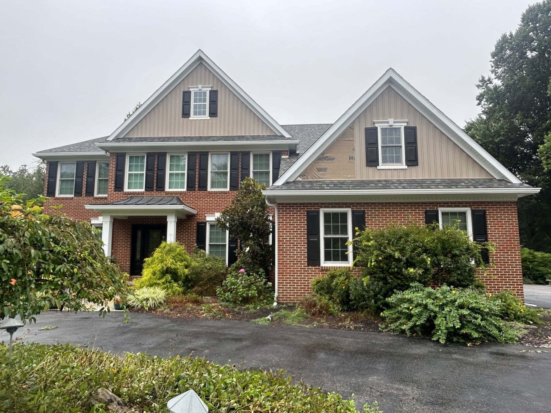 Two-story brick house with brown shingles, black shutters, and a driveway with overgrown bushes.
