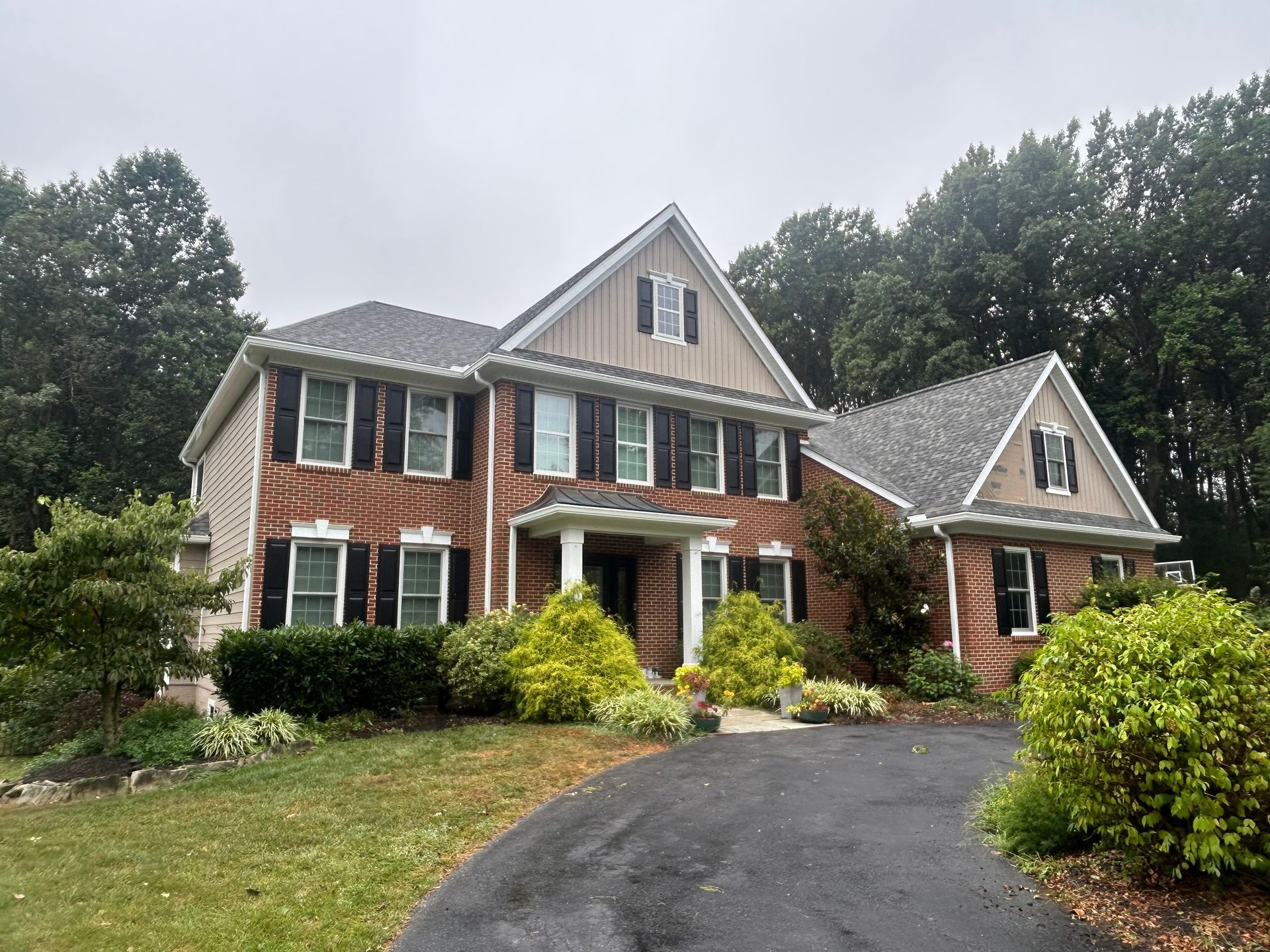 Two-story brick house with black shutters, driveway, and surrounding trees on an overcast day.