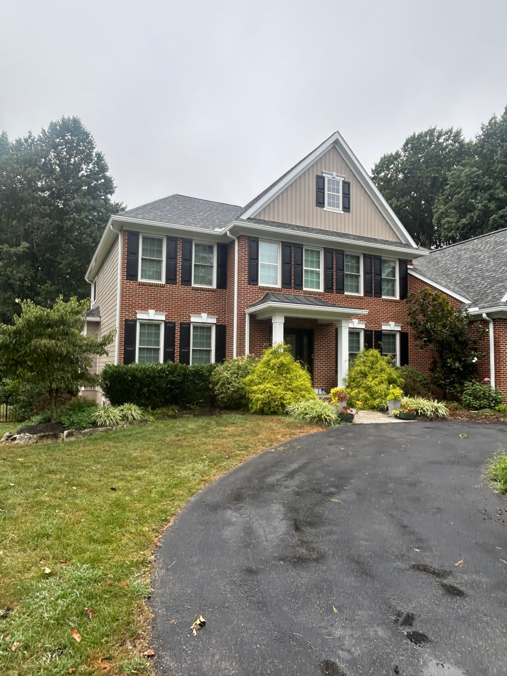 Two-story brick house with black shutters and a dark driveway, surrounded by trees and landscaping.
