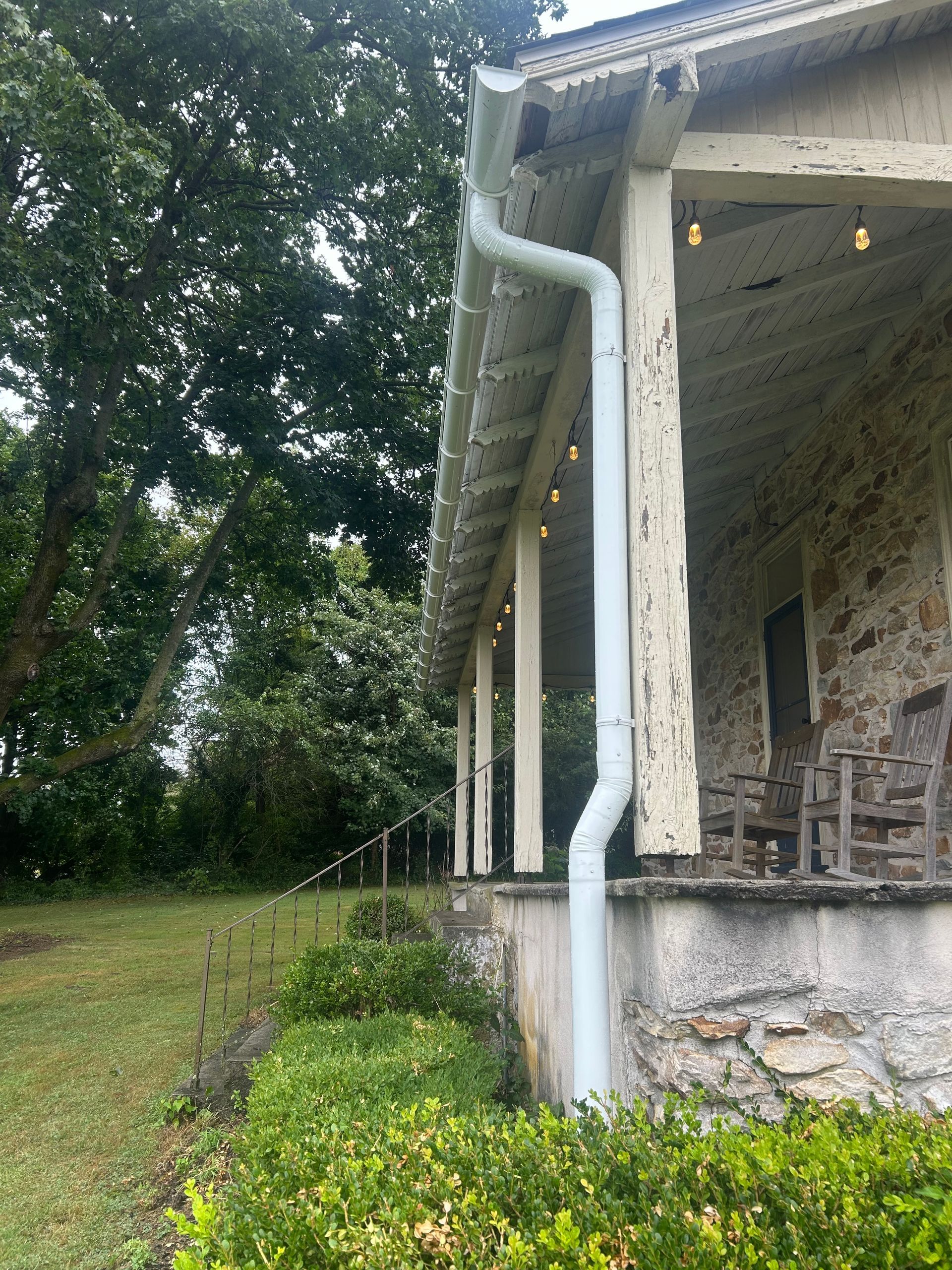 White gutter downspout on the corner of a stone building with a porch; green grass and foliage in the foreground.