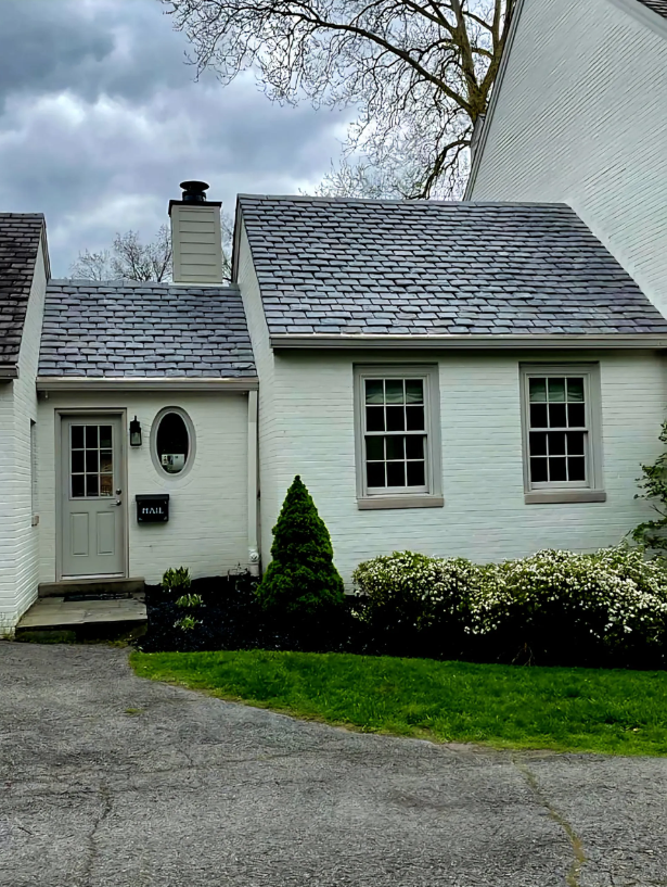 Small white house with gray shingled roof, front door, windows, and a driveway under cloudy sky