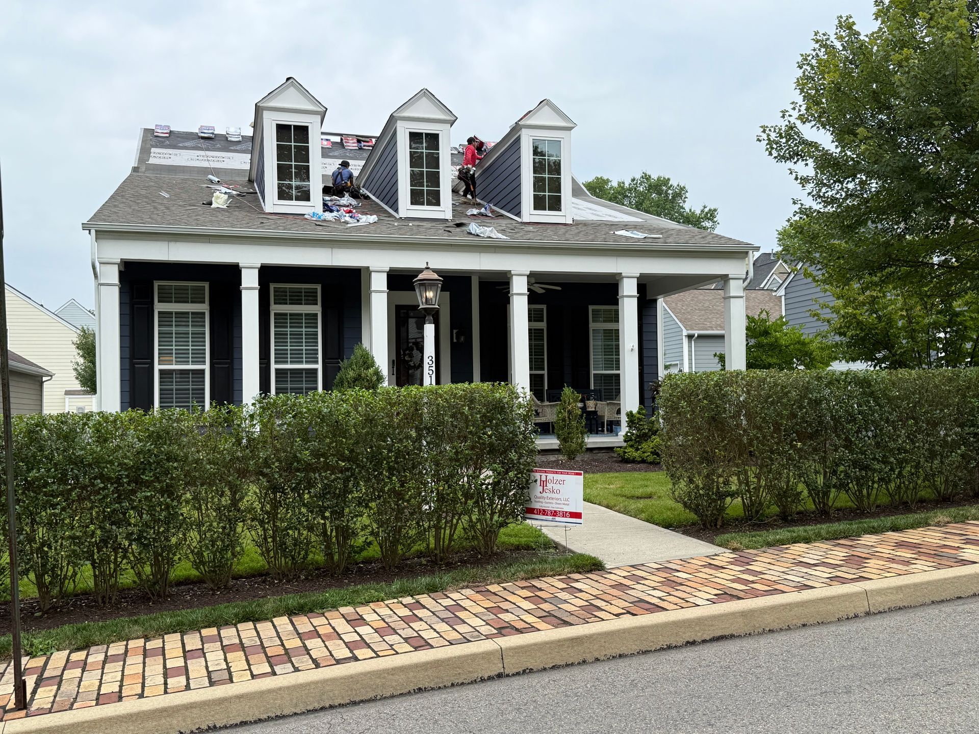 Black house with white trim, dormer windows, and a manicured hedge along the sidewalk