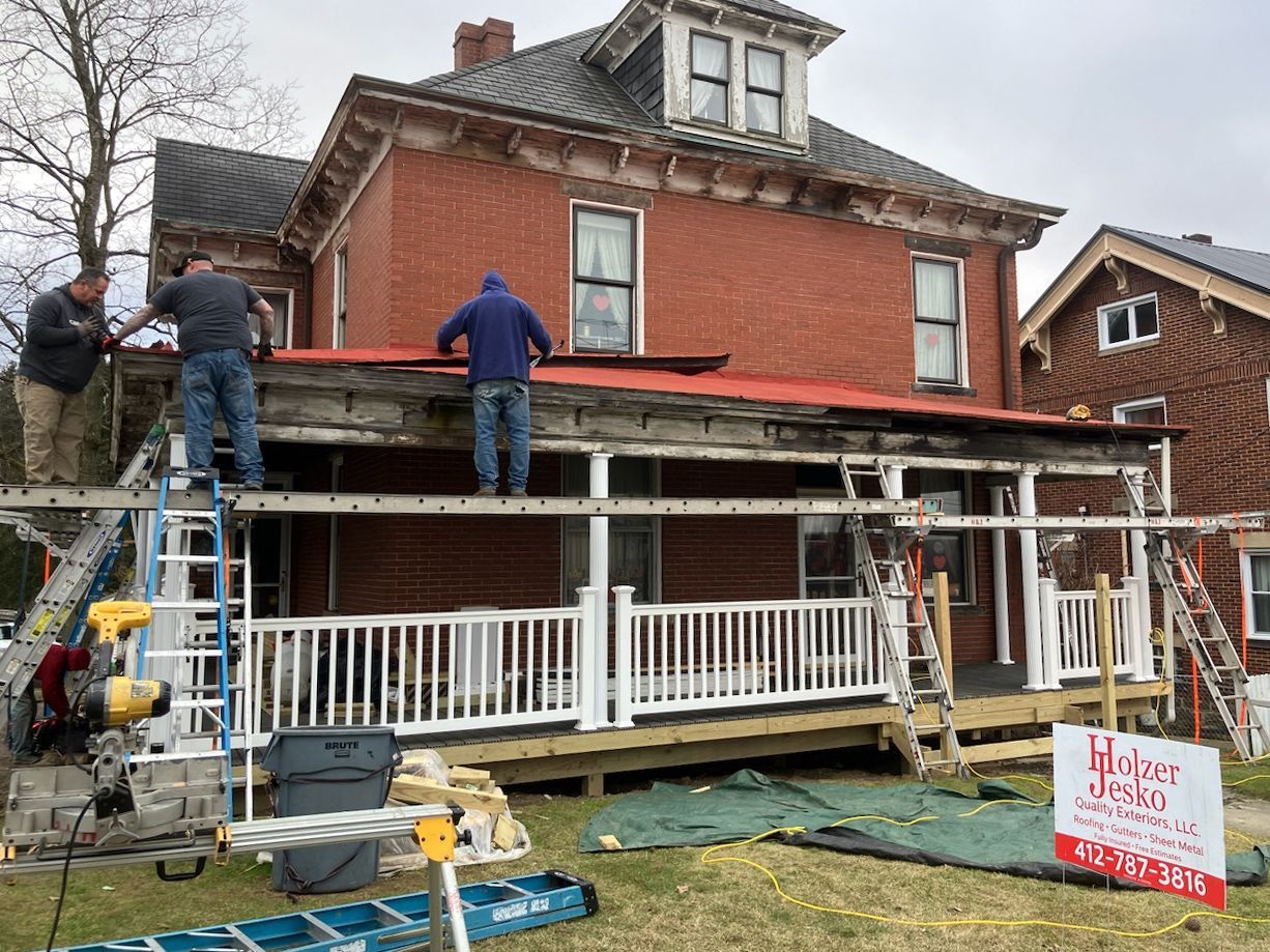 Workers repairing a red brick house porch roof with scaffolding and a ladder in front.