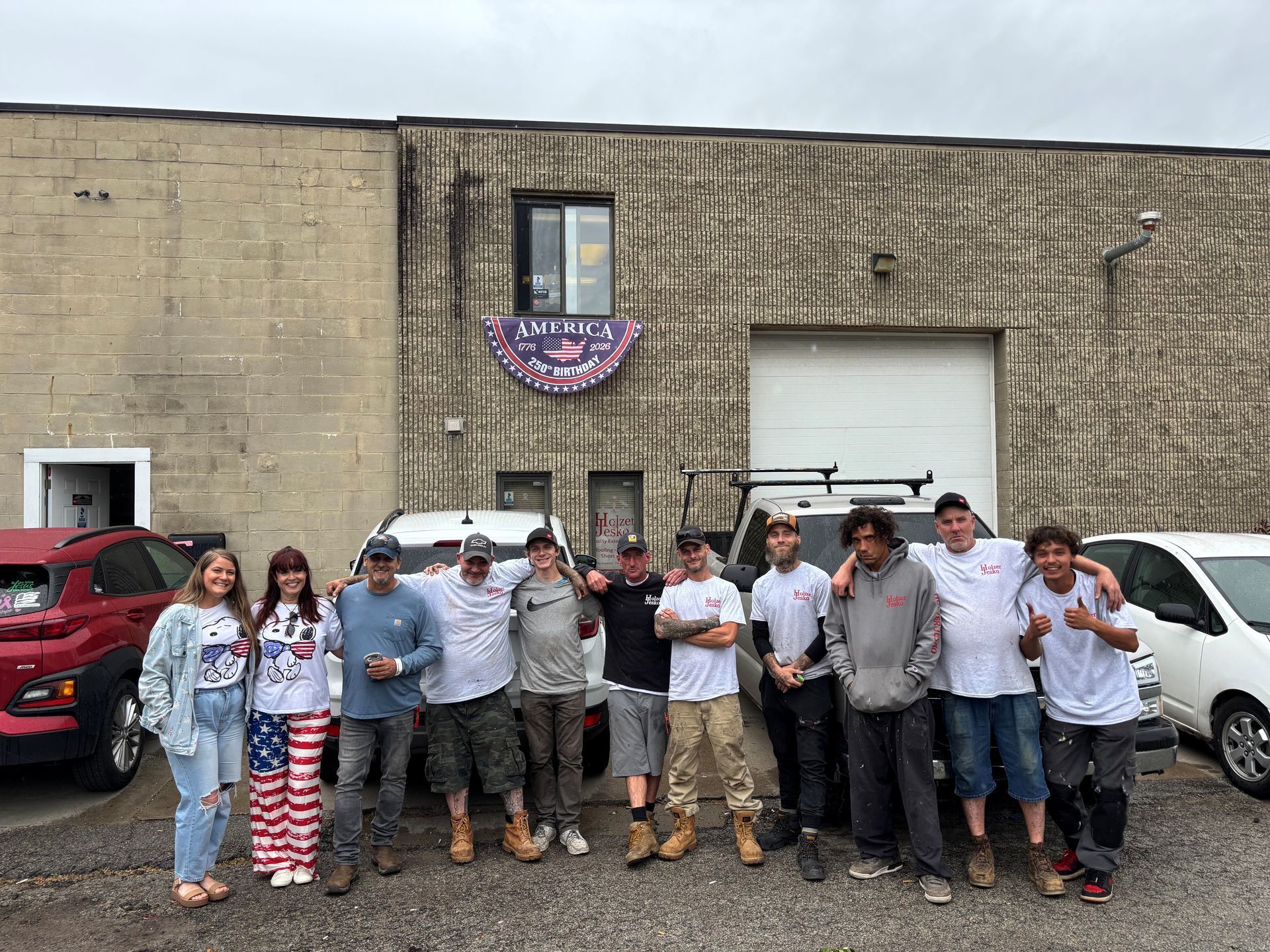 Group of people standing together outside a warehouse with parked vehicles, posing for a photo