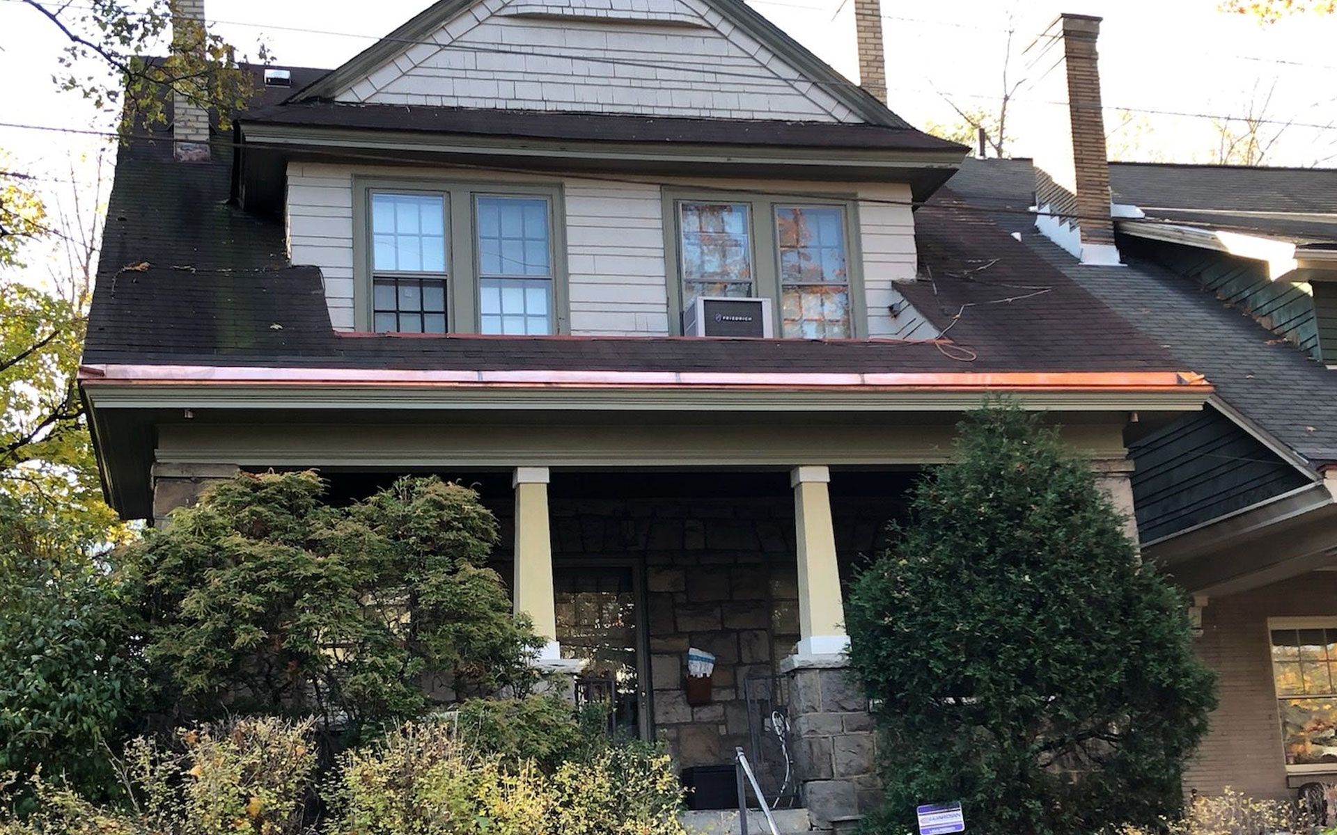 Old two-story house with a porch, dark roof, and overgrown shrubs in front