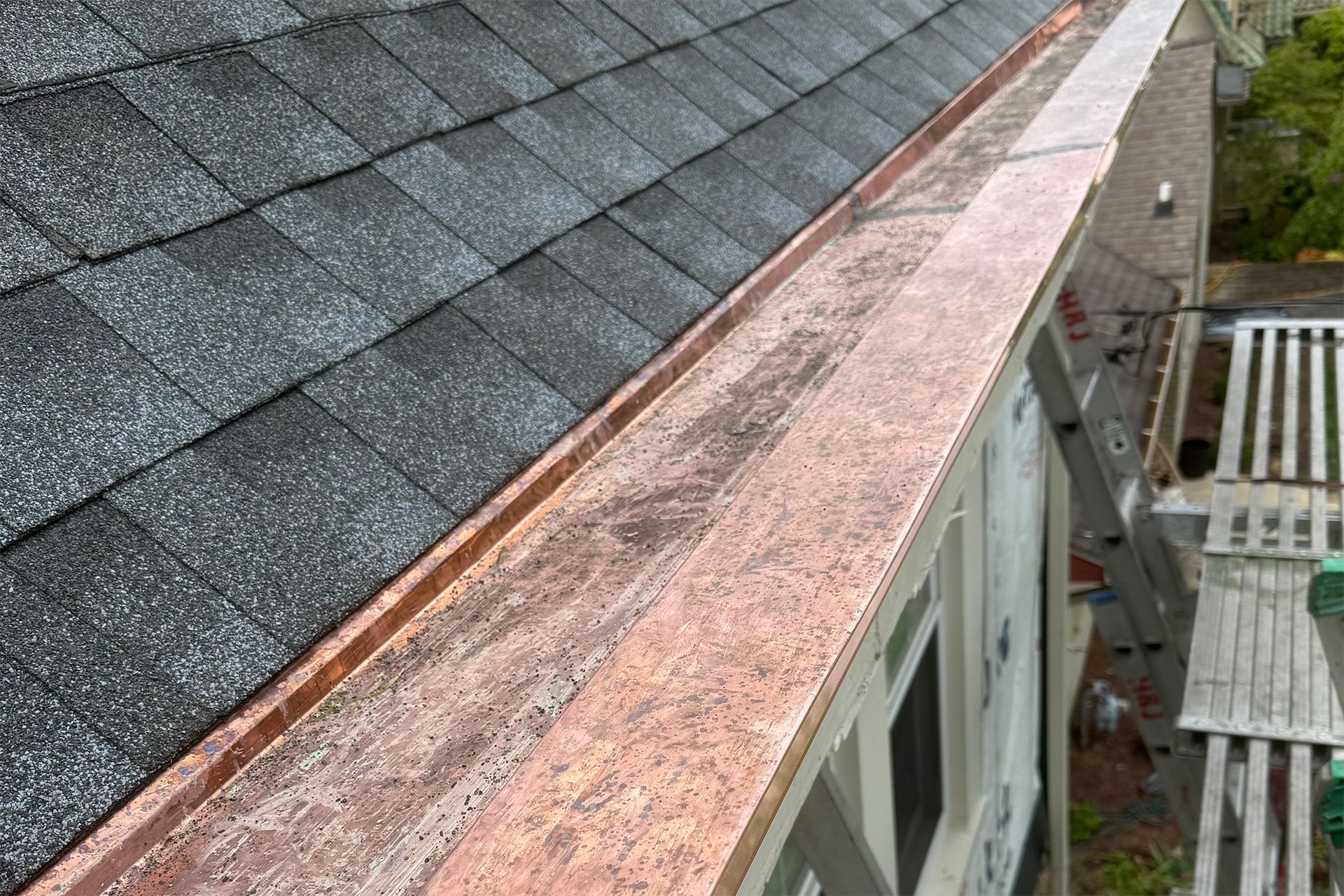 Roof edge with dark shingles and a rusty copper gutter beside a house and patio below