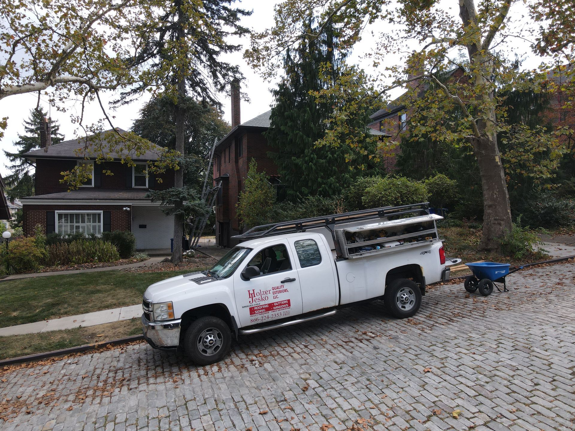 White utility truck parked on a cobblestone street beside a small trailer, with houses and trees in the background