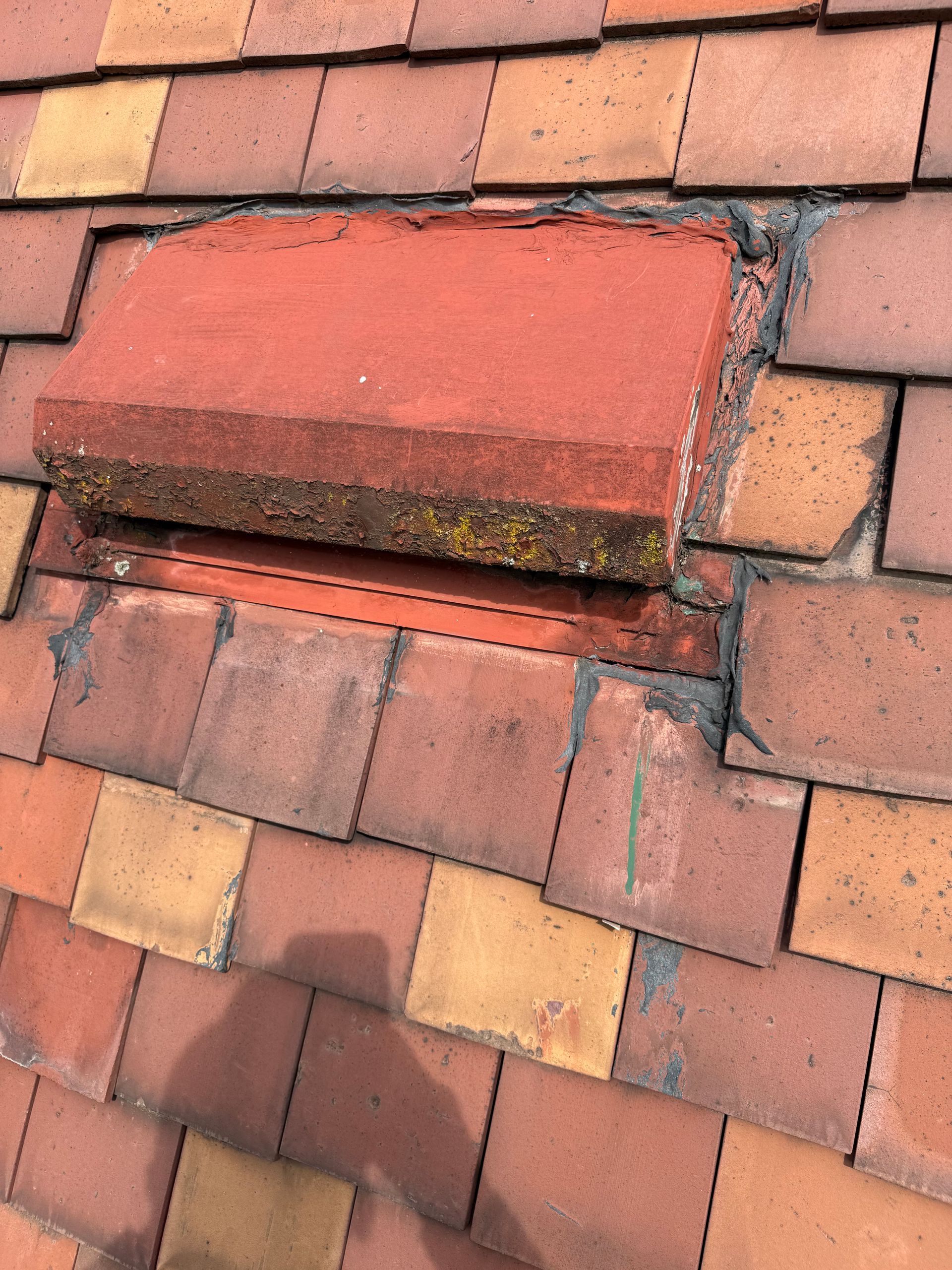 Damaged red roof vent among weathered terracotta tiles, with moss and cracked mortar