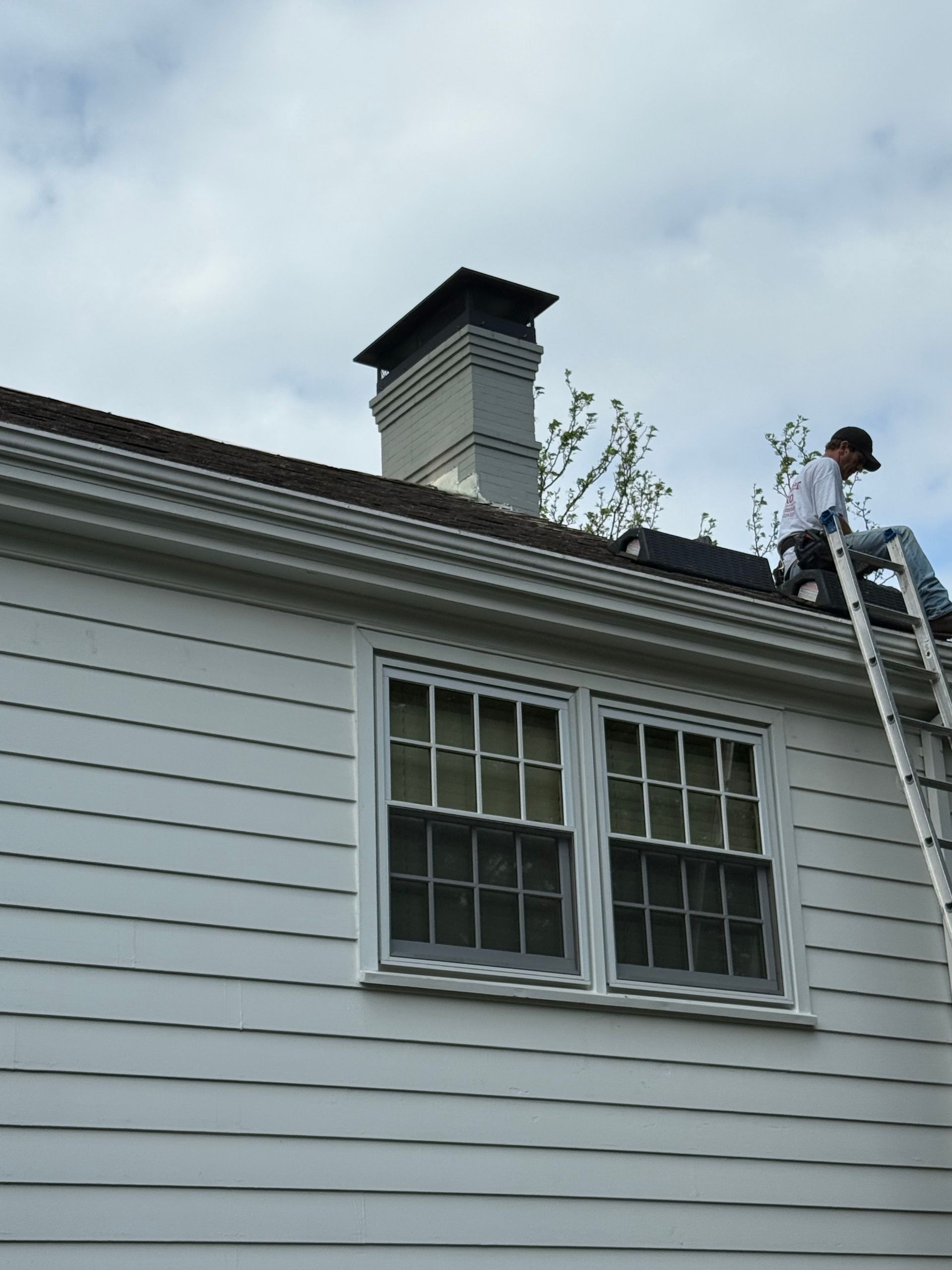 Worker on a ladder cleaning a white house roof near a chimney