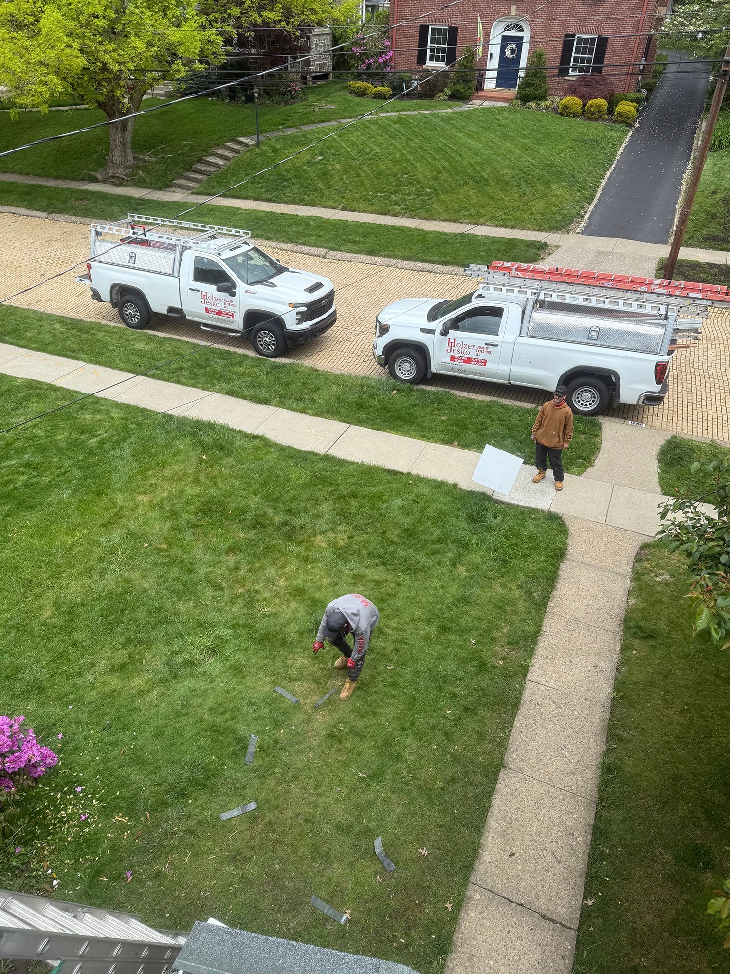 Two white utility vans parked beside a brick house, with two workers on a grassy lawn and sidewalk.