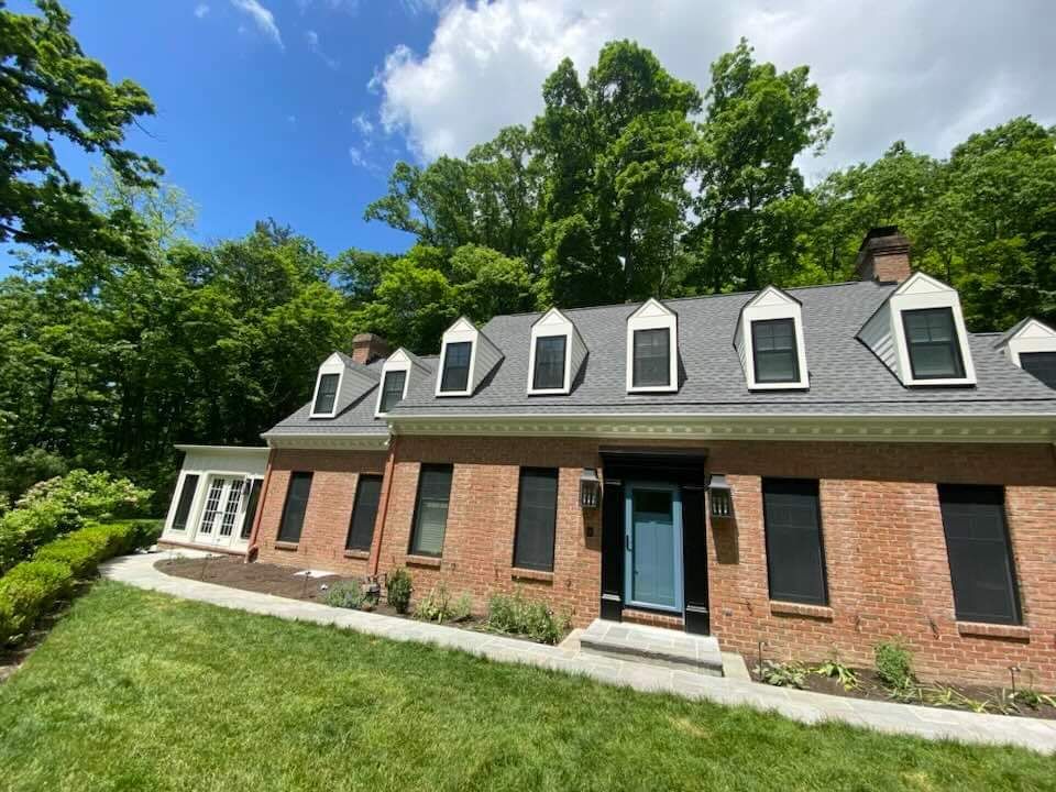Brick house with dormer windows and black shutters, set in a green wooded yard under a blue sky