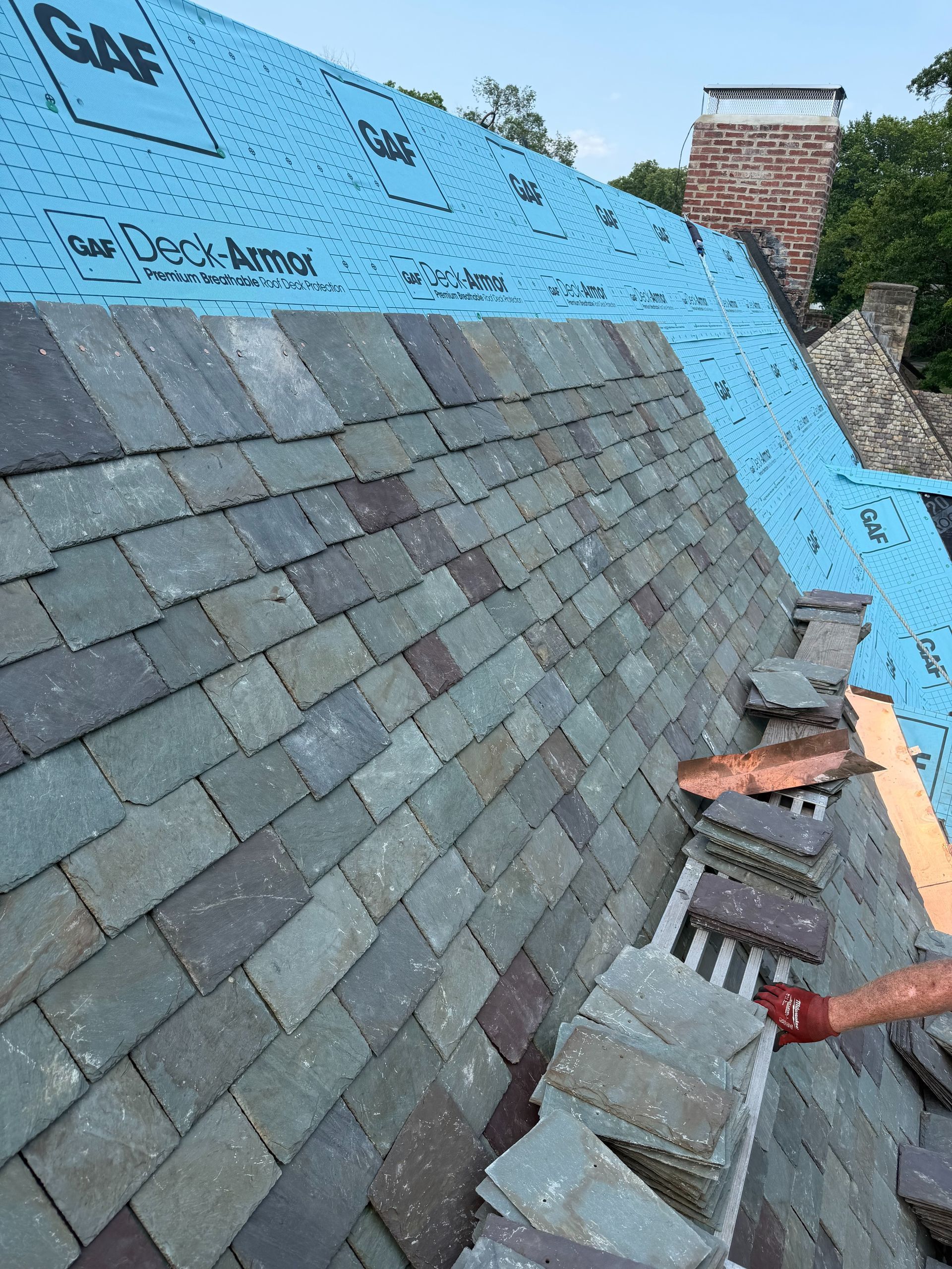 Roof under repair with blue underlayment and stacked shingles beside a chimney