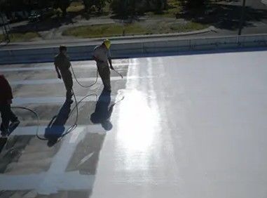 Workers spraying white coating on a large rooftop, creating reflective surface. Sunny conditions.