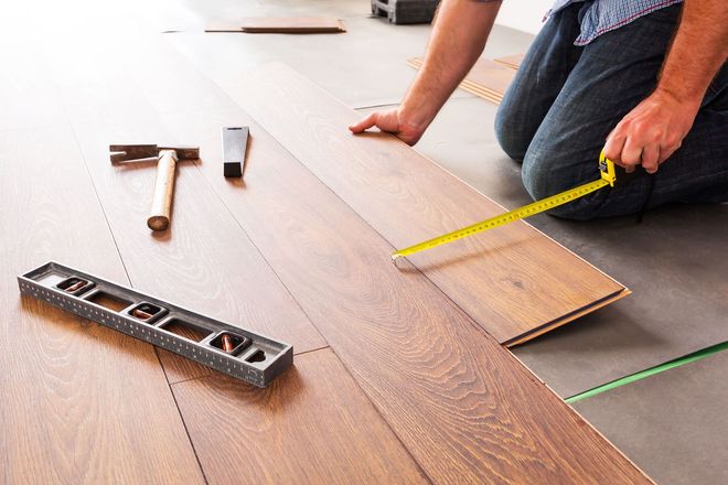 Person installing hardwood flooring, measuring a board with a tape measure and tools nearby.