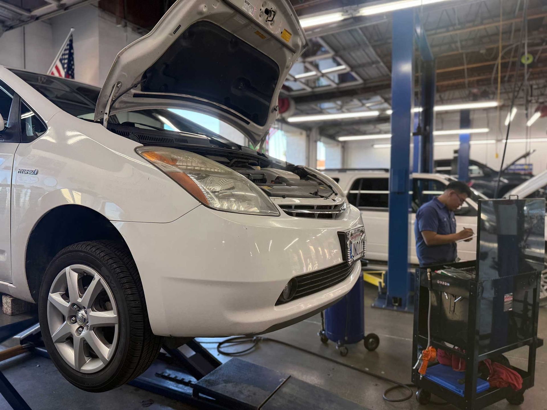 White car on a lift in a repair shop with its hood open; a mechanic works nearby.