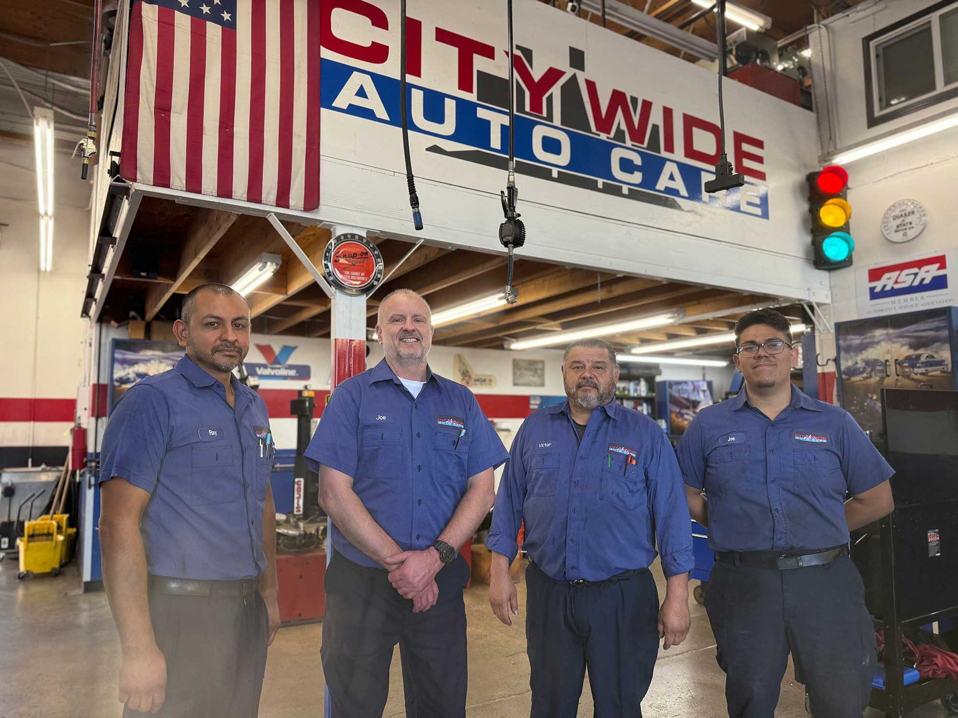 Four auto mechanics in blue uniforms standing inside City Wide Auto Care. An American flag and logo are visible.