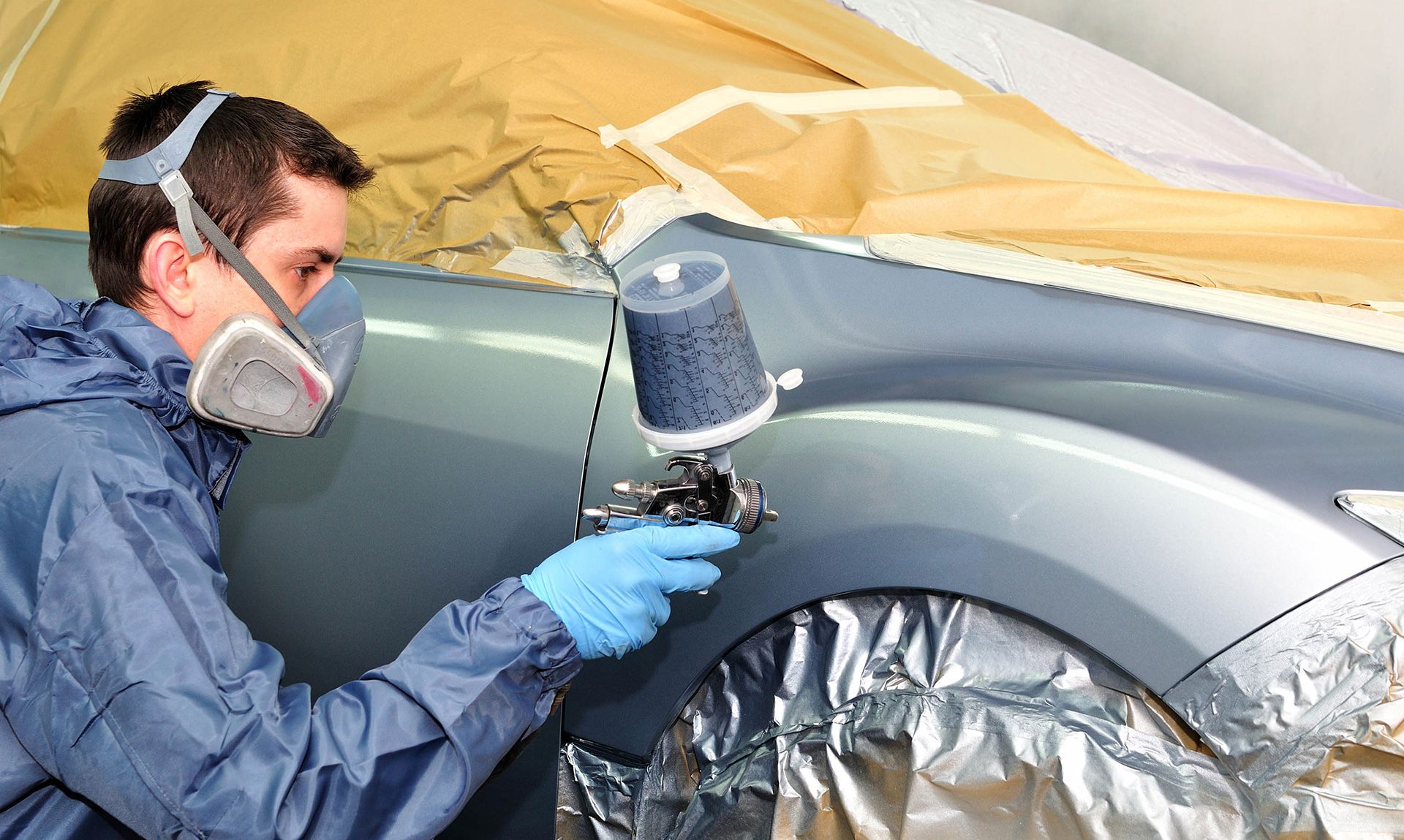 A person in protective gear sprays paint on a car fender, masking tape and paper visible.