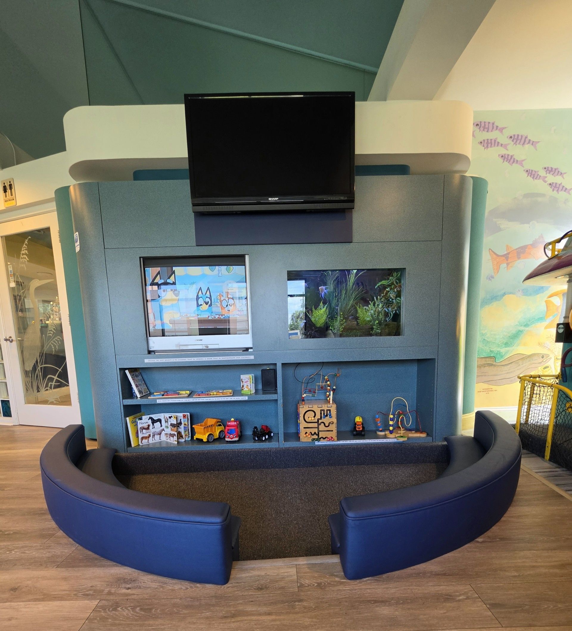 Blue aquarium-themed booth with a TV on top and shelves of colorful books and toys, framed by a yellow seat