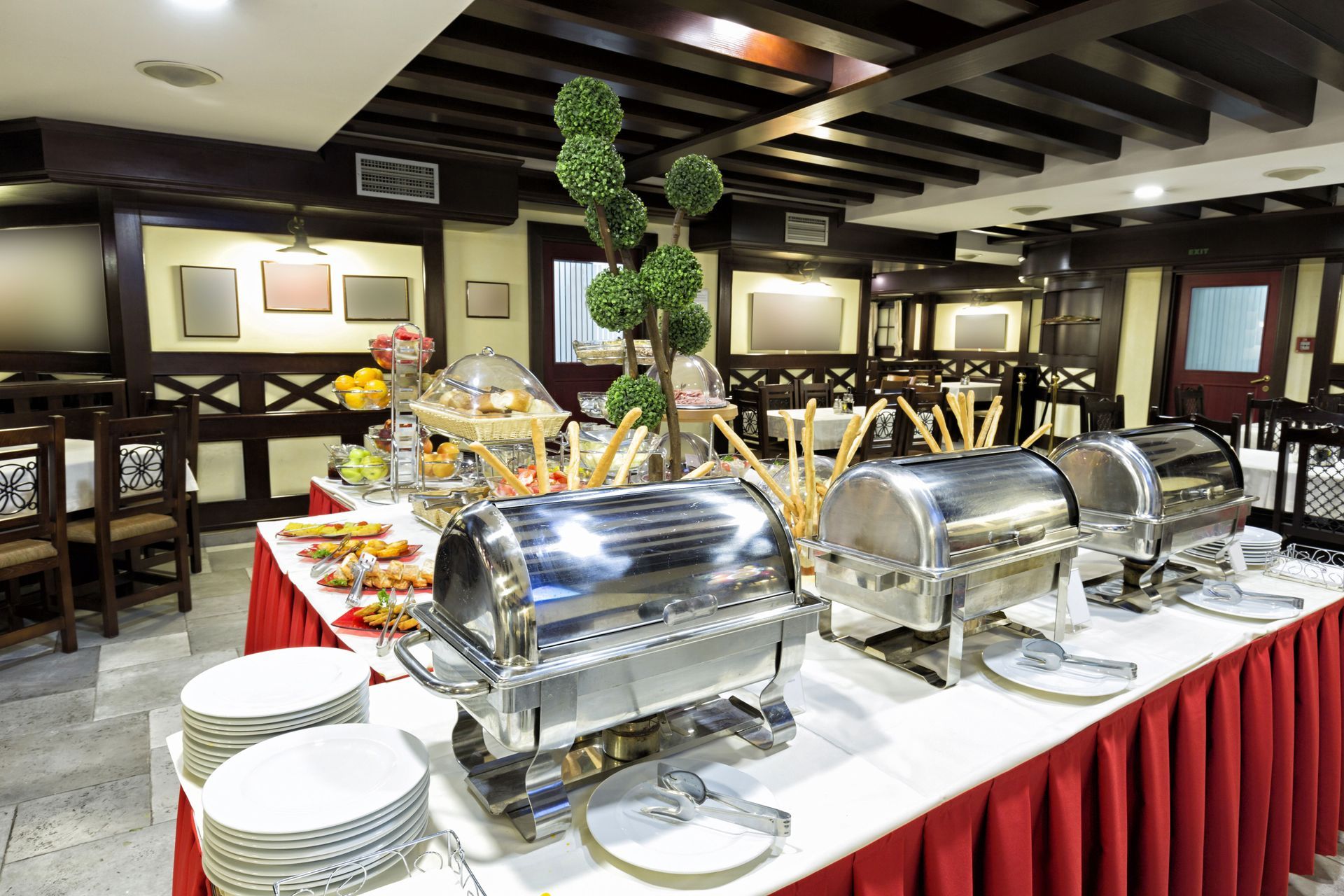 Buffet line with covered chafing dishes, stacked plates, and food trays in a restaurant dining room