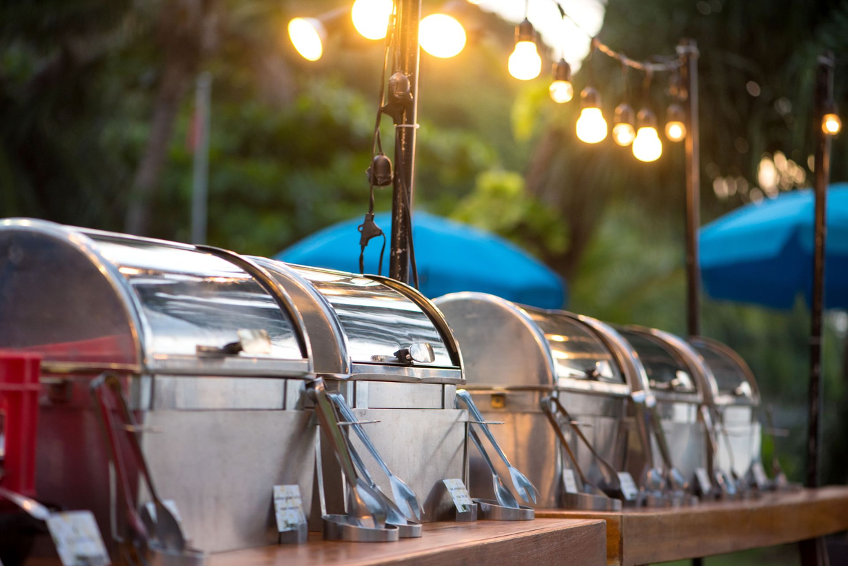 Row of stainless steel buffet chafing dishes on a wooden table under warm outdoor string lights