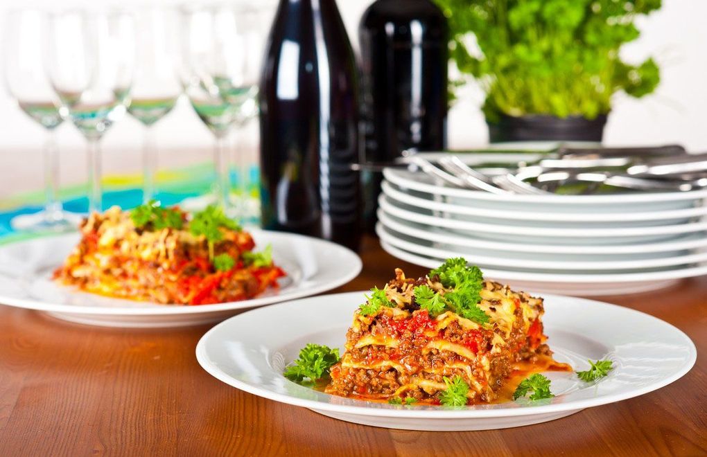 Plated pasta dish on a wooden table with wine glasses, bottles, and stacked plates in the background