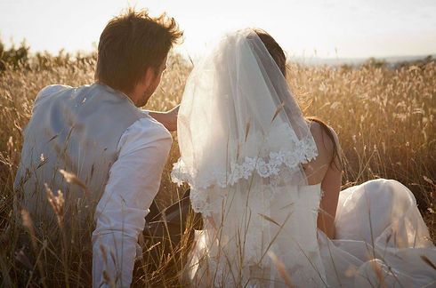 Couple in wedding attire sitting in tall grass at sunset, facing each other