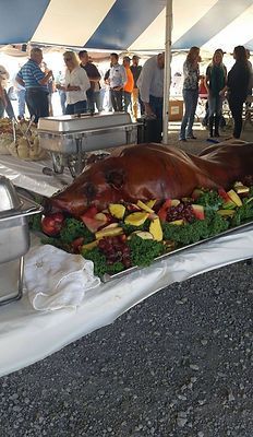 Roasted pig on a buffet table with vegetables under a striped event tent, people standing nearby