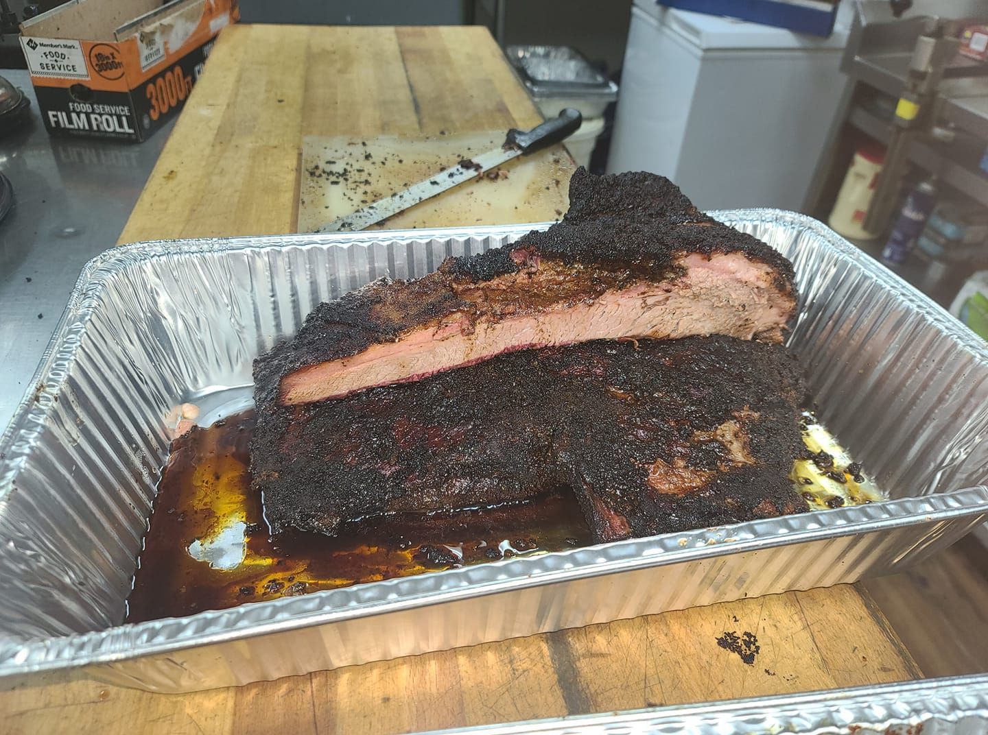 Smoked brisket in a foil pan on a wooden table, with a sliced section showing a dark bark and pink smoke ring