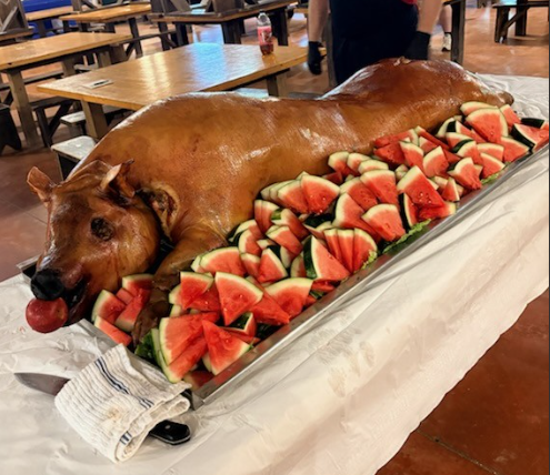 Whole roasted pig on a table, surrounded by sliced watermelon in an outdoor dining area