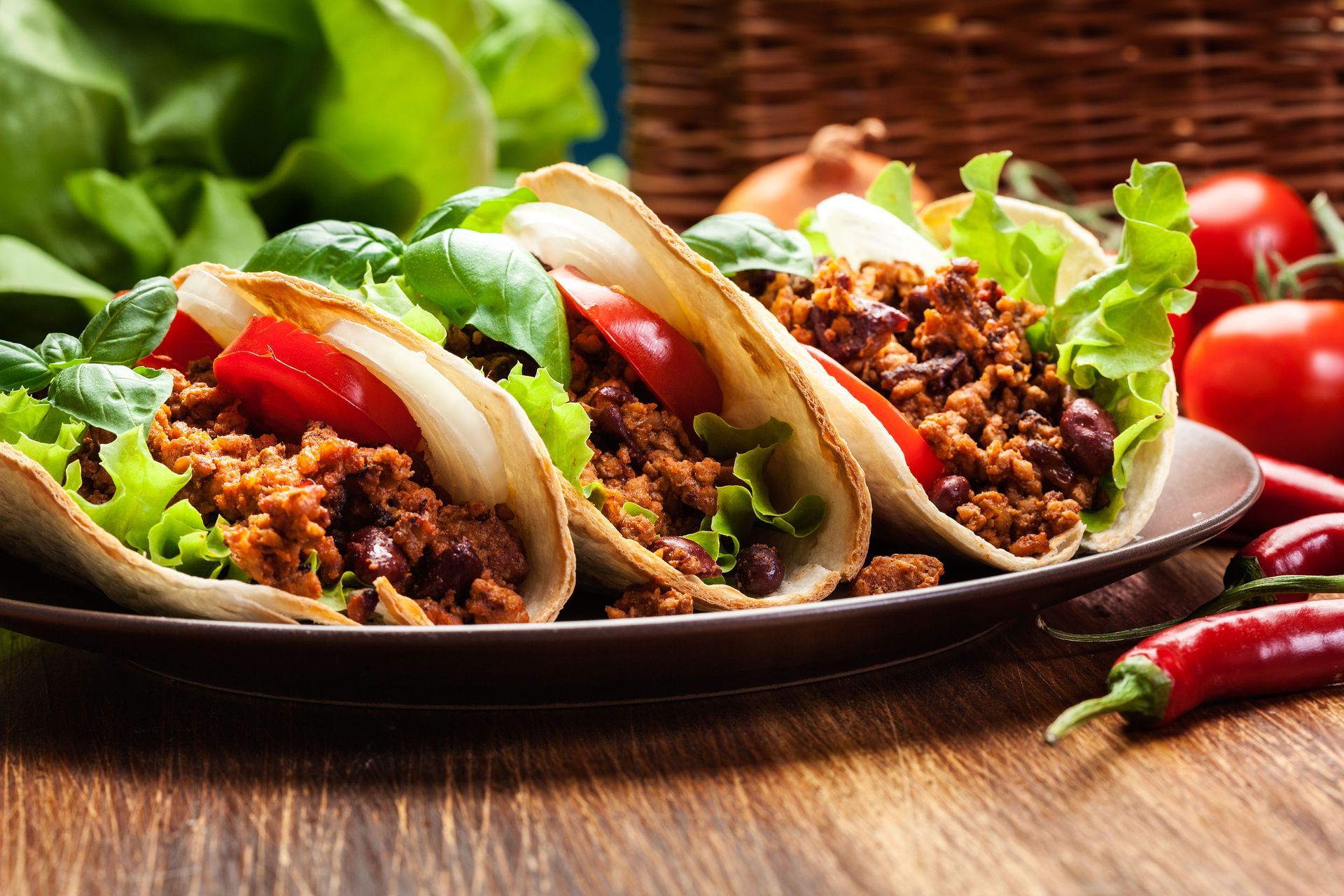Tacos with beef, lettuce, tomato, and cilantro on a dark plate, with chilies on a wooden table.