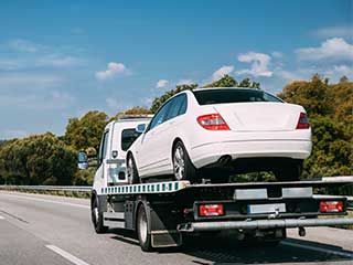 a white car being towed on a flatbed truck on a highway, under a blue sky with trees