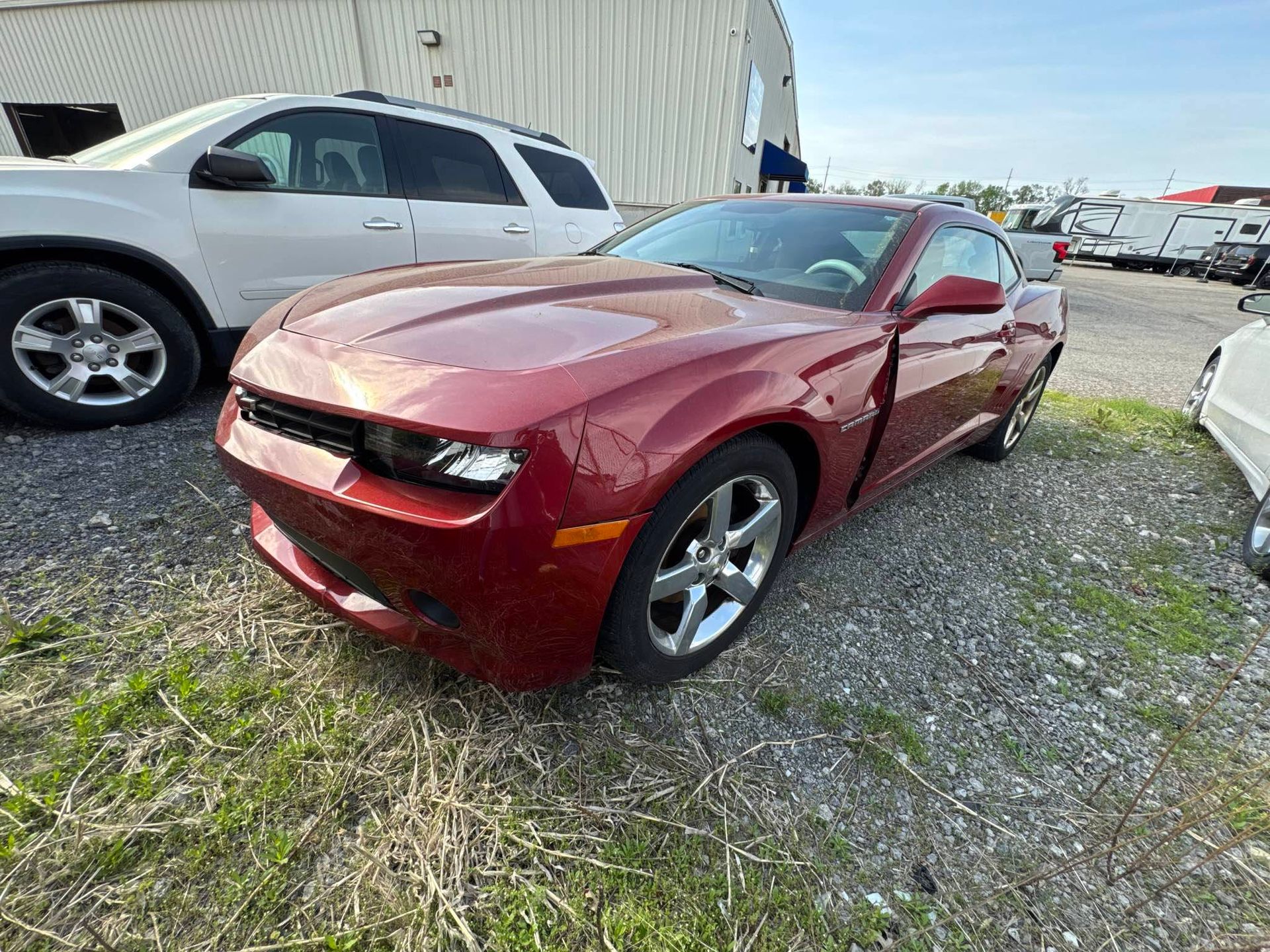 Red sports car with front-end damage parked on gravel beside a white SUV and warehouse.
