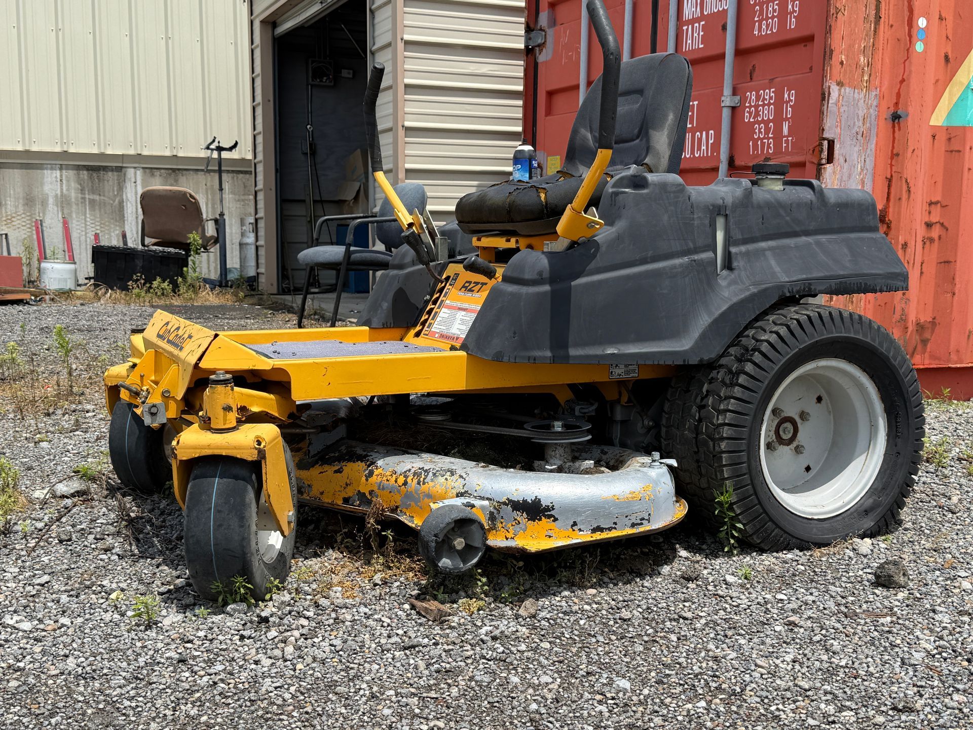 Yellow riding lawn mower in a gravel lot beside a building and shipping containers