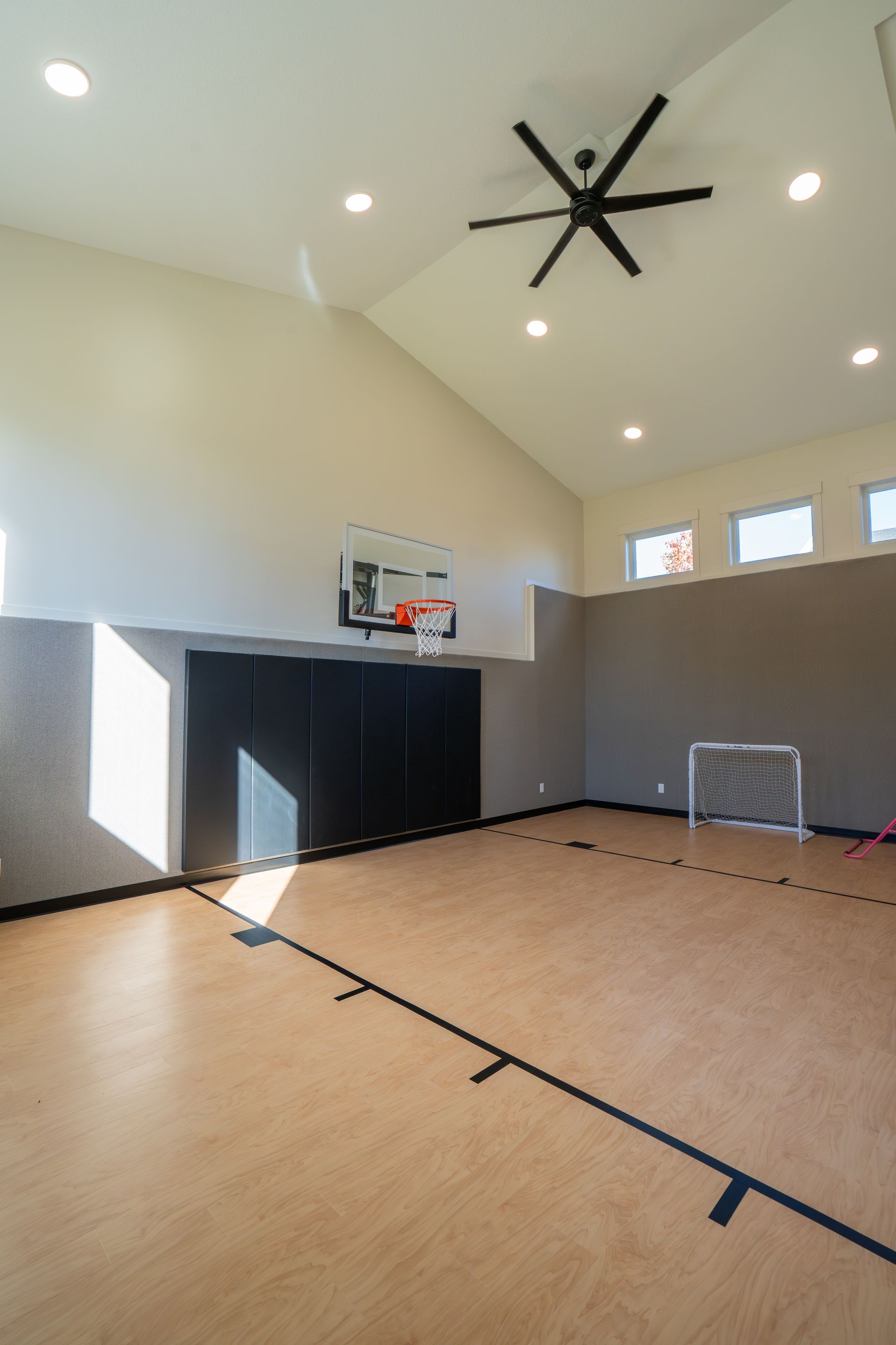 Indoor basketball court with a hoop, high ceiling fan, and bright windows.