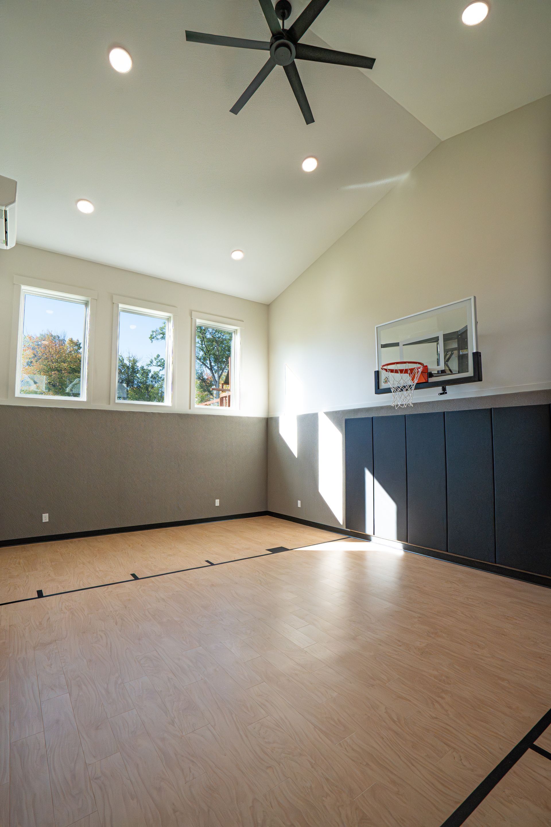 Empty indoor basketball half-court with high ceiling, black fans, windows, and wall padding