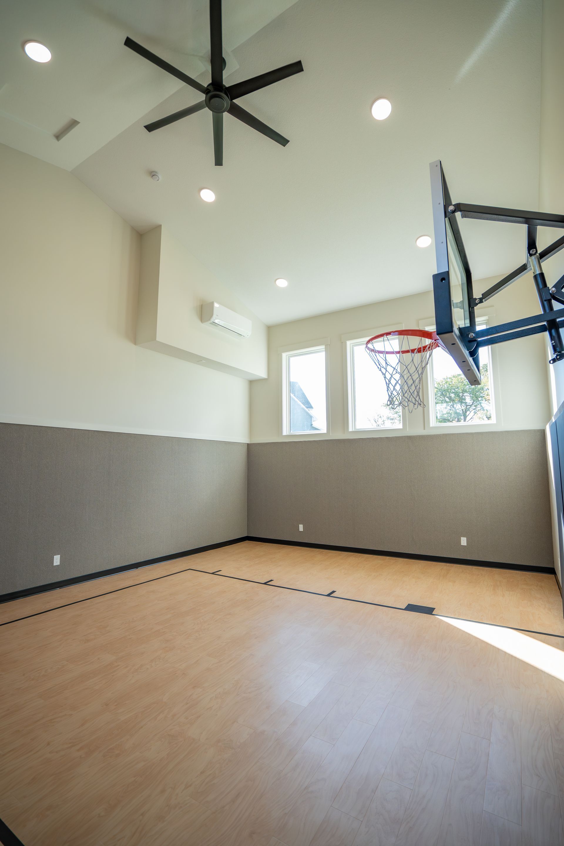Indoor basketball court with wood floor, hoop, high ceiling fan, and windows letting in bright daylight