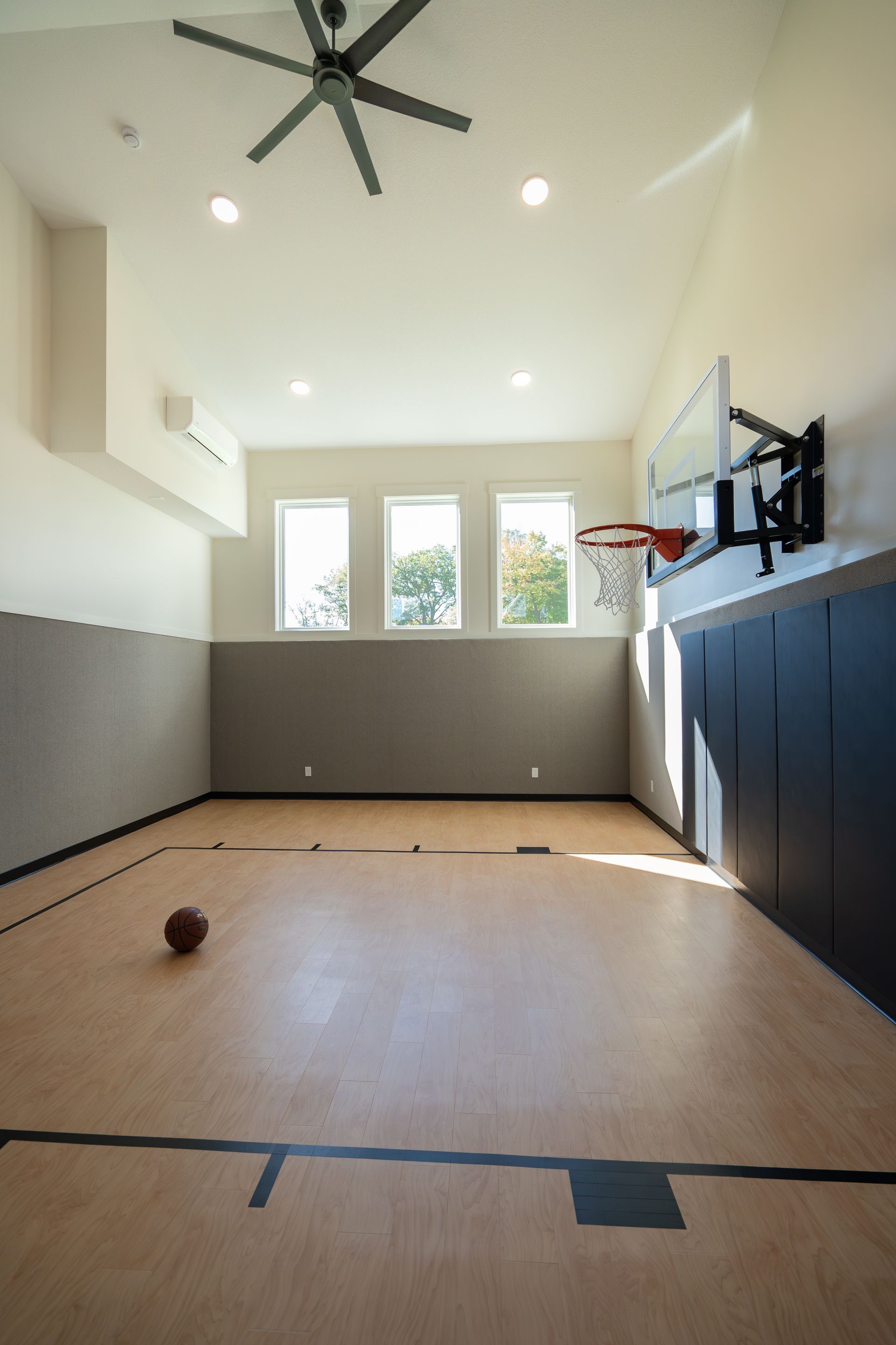Indoor basketball court with hardwood floor, hoop, windows, and a ceiling fan above.