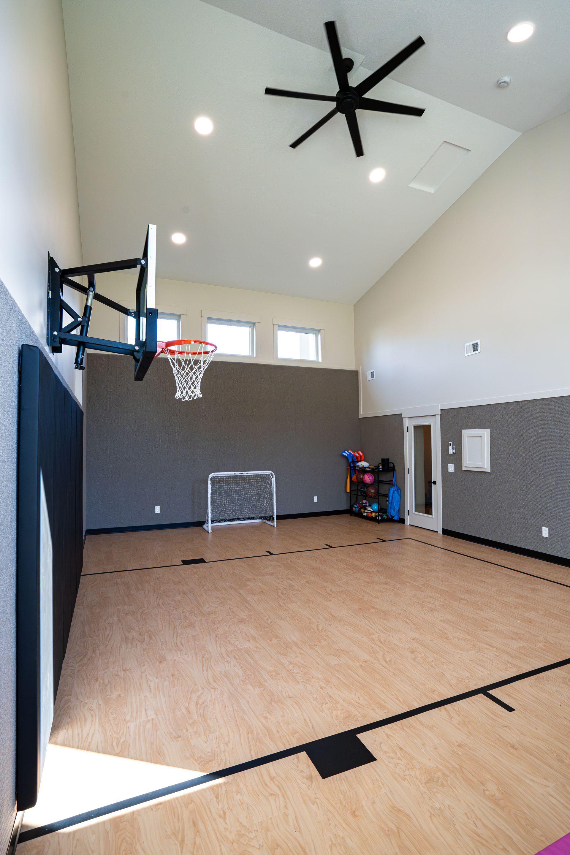 Indoor basketball court with hoop, high ceiling fan, gray walls, and wood floor