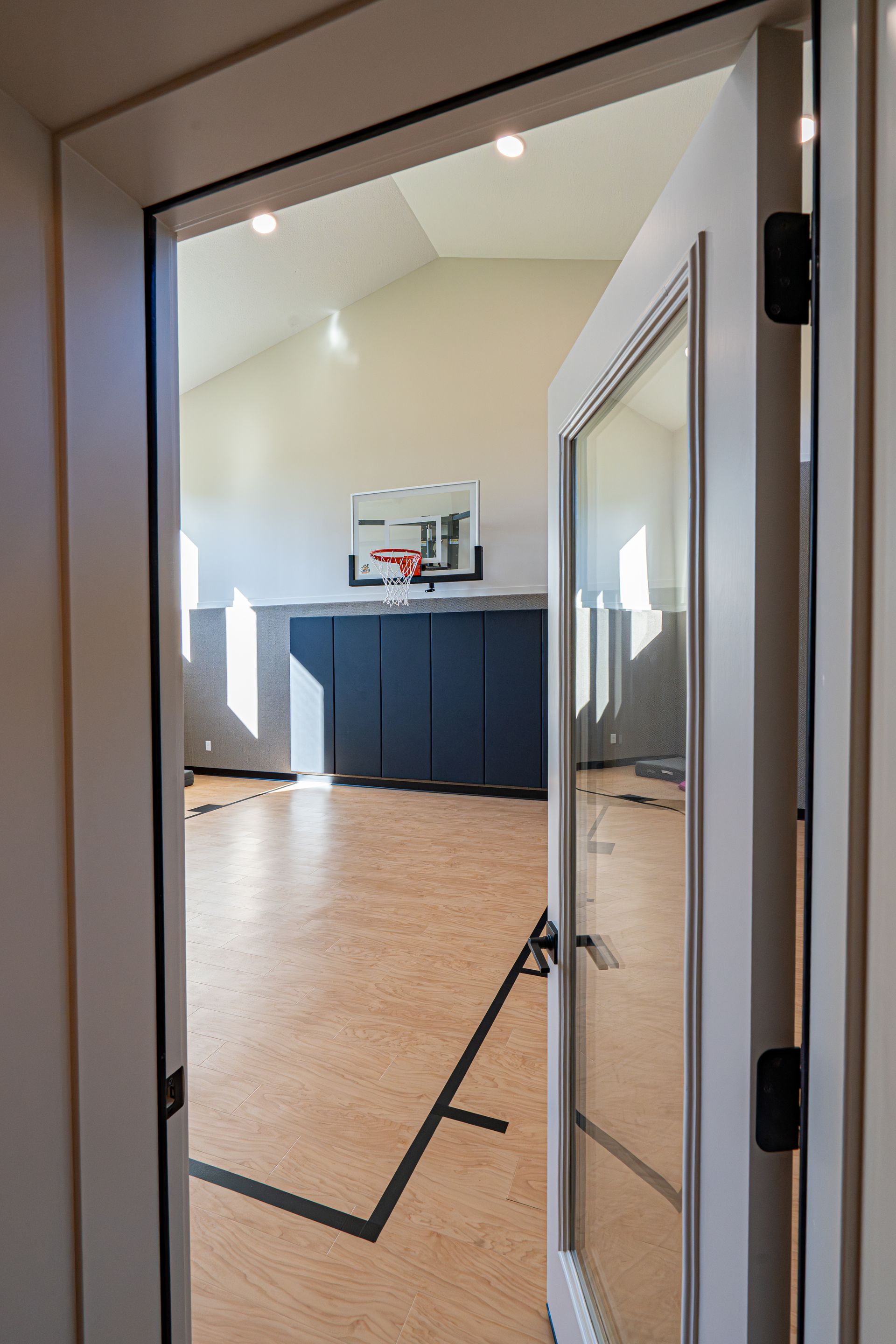 Empty gym seen through a doorway, with a basketball hoop and dark padded wall panels.