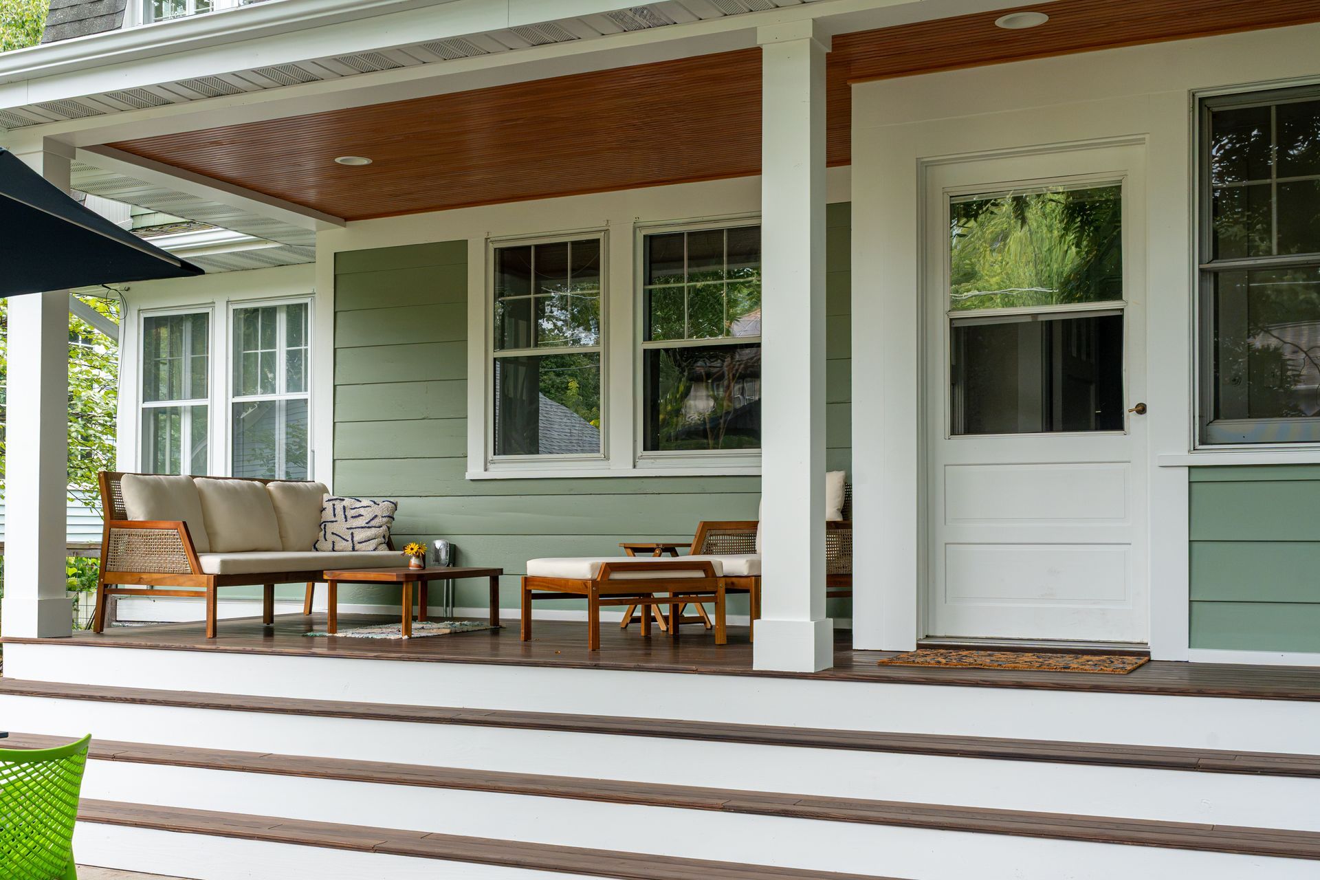 Covered porch with green siding, white columns, and a wicker bench, chairs, and table.