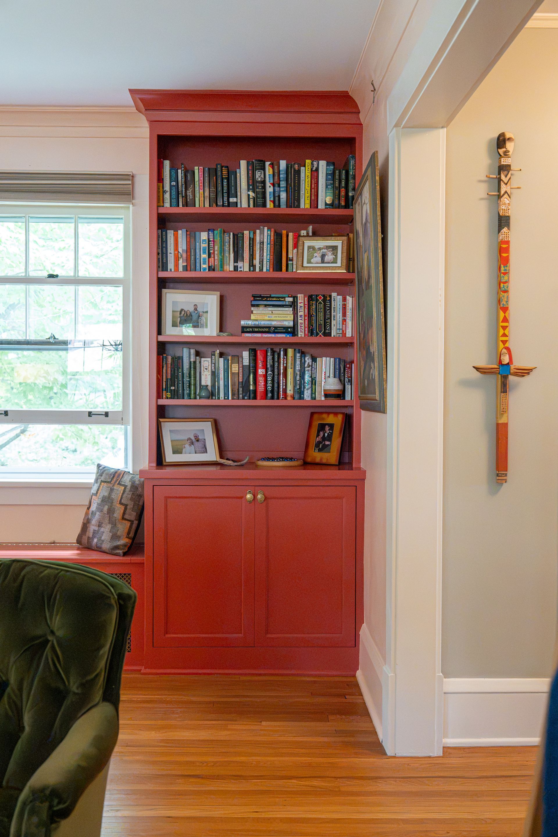 Built-in red bookshelf filled with books beside a wall-mounted decorative sword in a bright room