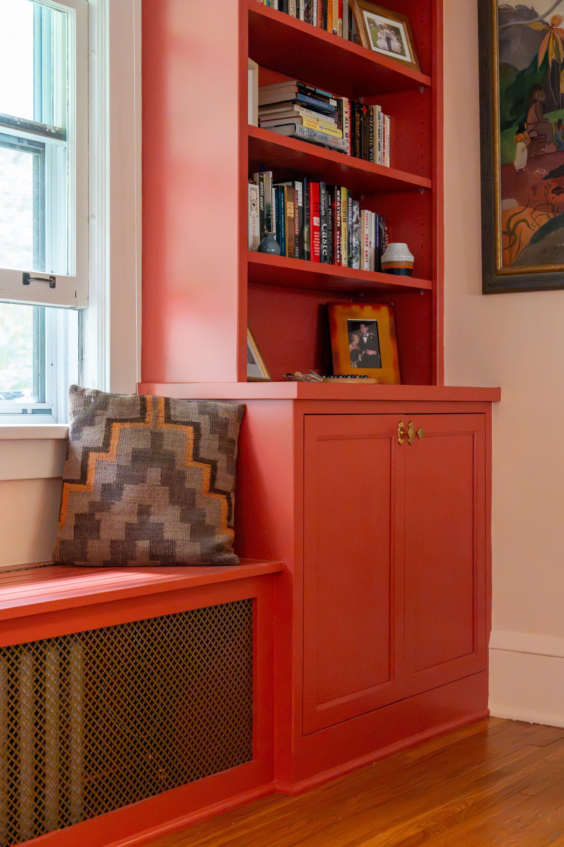 Red built-in bookshelf and cabinet beside a window, with books and a patterned cushion in a cozy room.