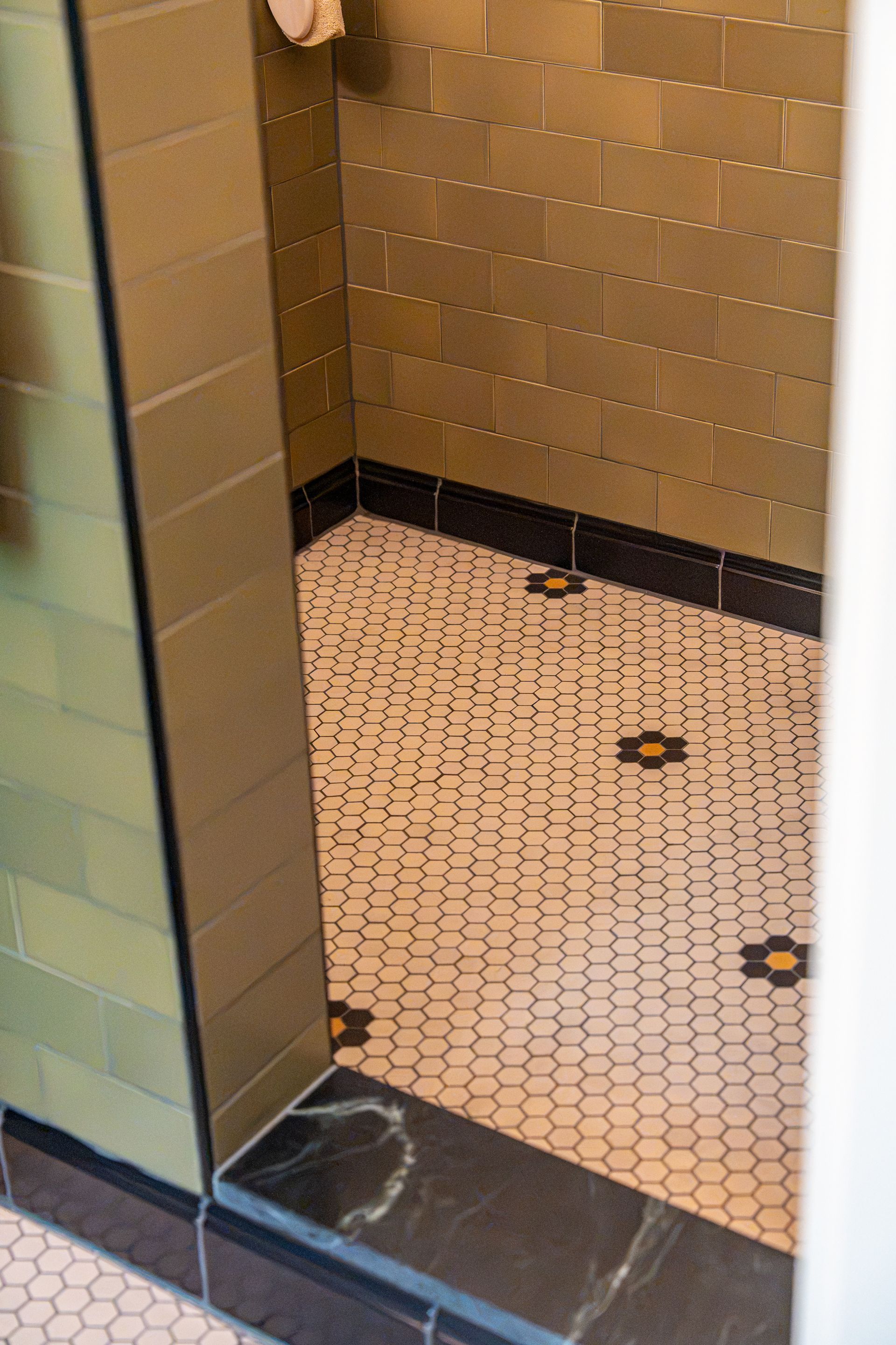 Public restroom shower stall with tan tile walls, hex floor tiles, and floor drains