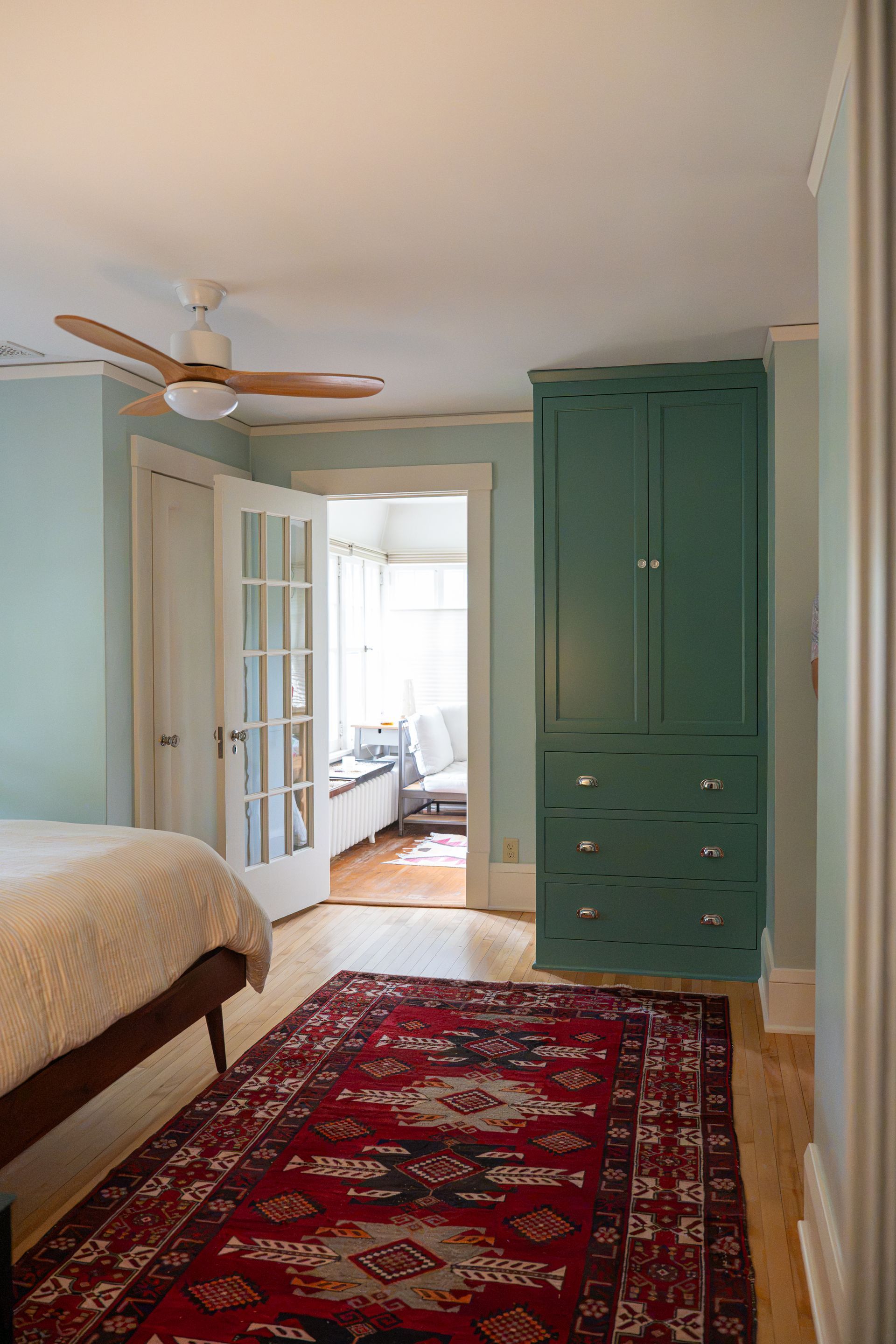 Bedroom with mint walls, red patterned rug, bed, ceiling fan, and green wardrobe by French doors.