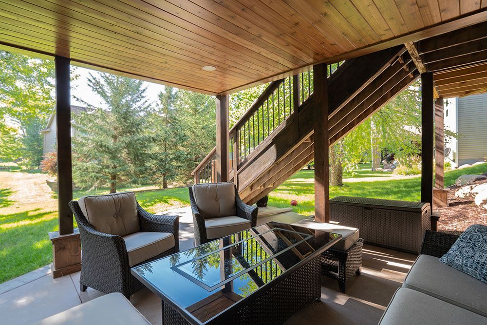 Covered patio seating area with wicker chairs, glass table, and stairs under a wooden roof.