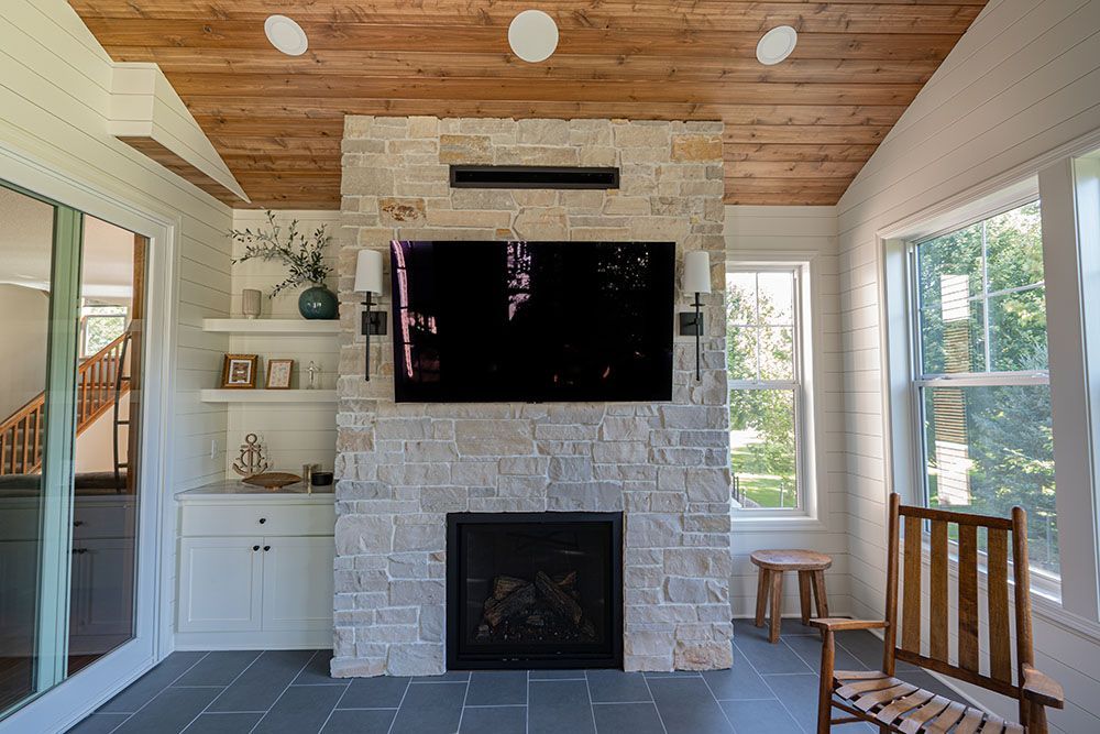 Bright living room with stone fireplace, mounted TV, white built-ins, and wood ceiling